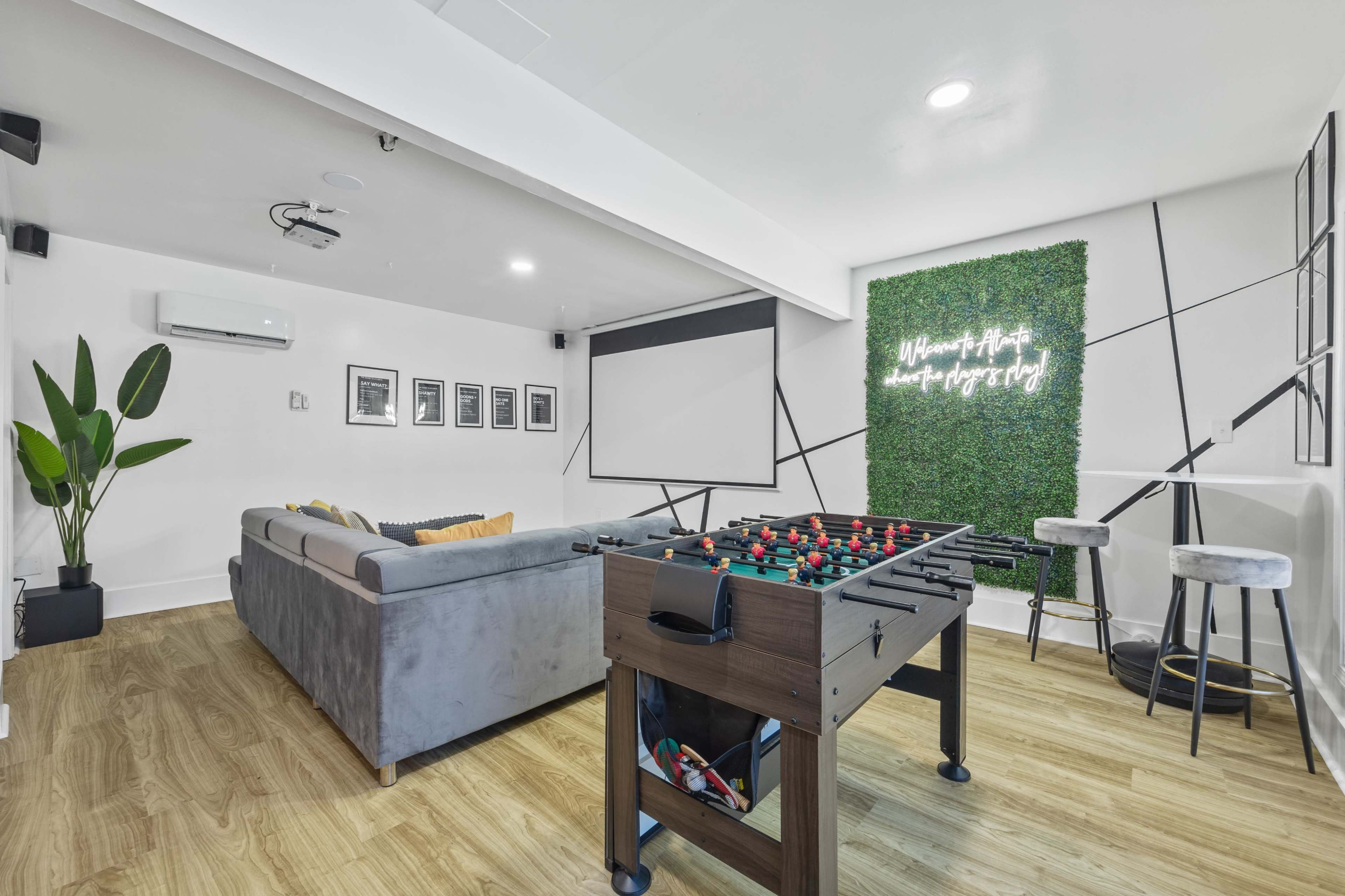 The image depicts a modern game room featuring a foosball table, a gray sectional sofa, a projector screen, and a green accent wall with decorative text.