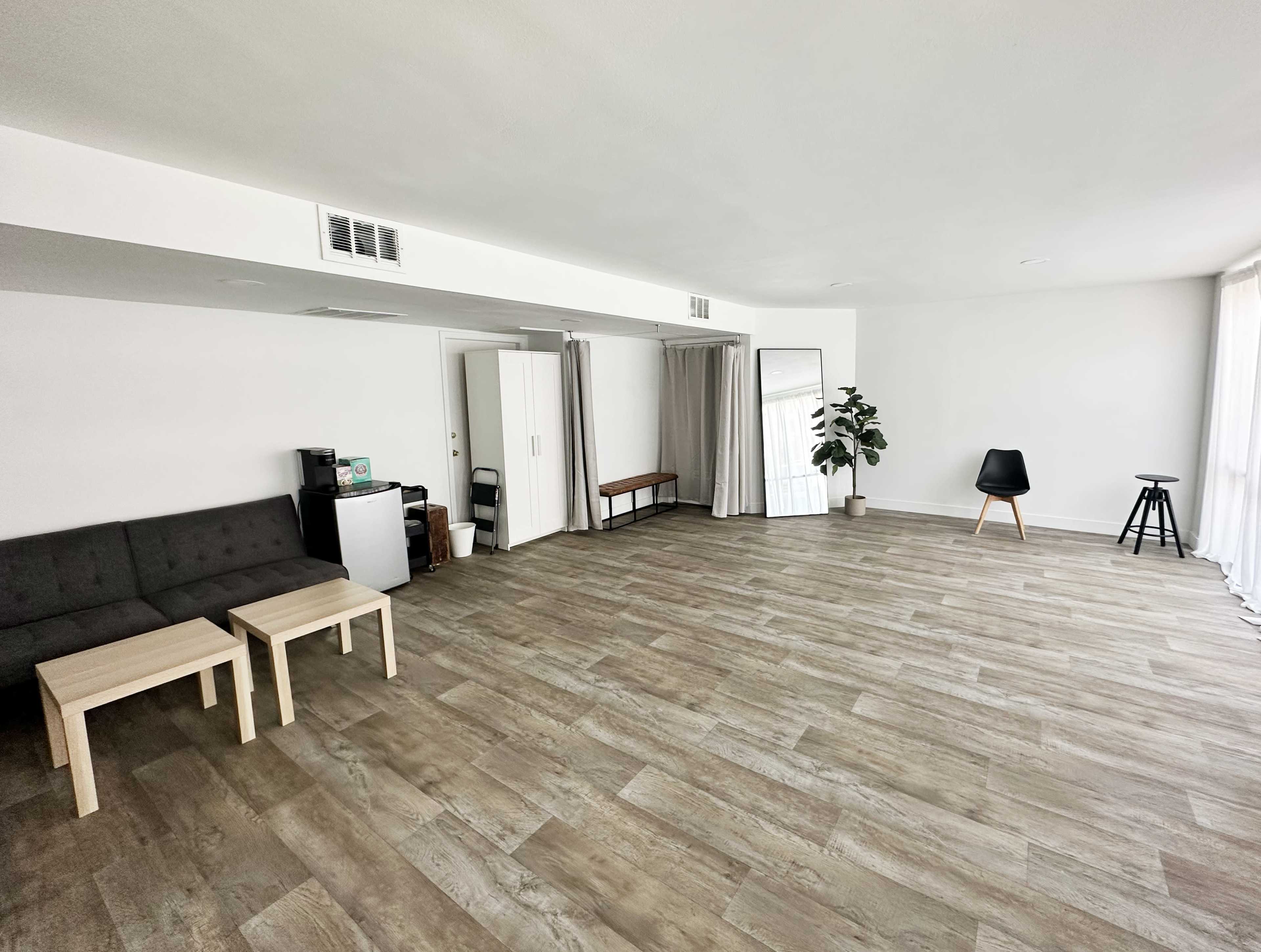 A spacious room with light wood flooring, a gray couch, two small tables, a chair, a mirror, and a plant next to a curtain-covered doorway.