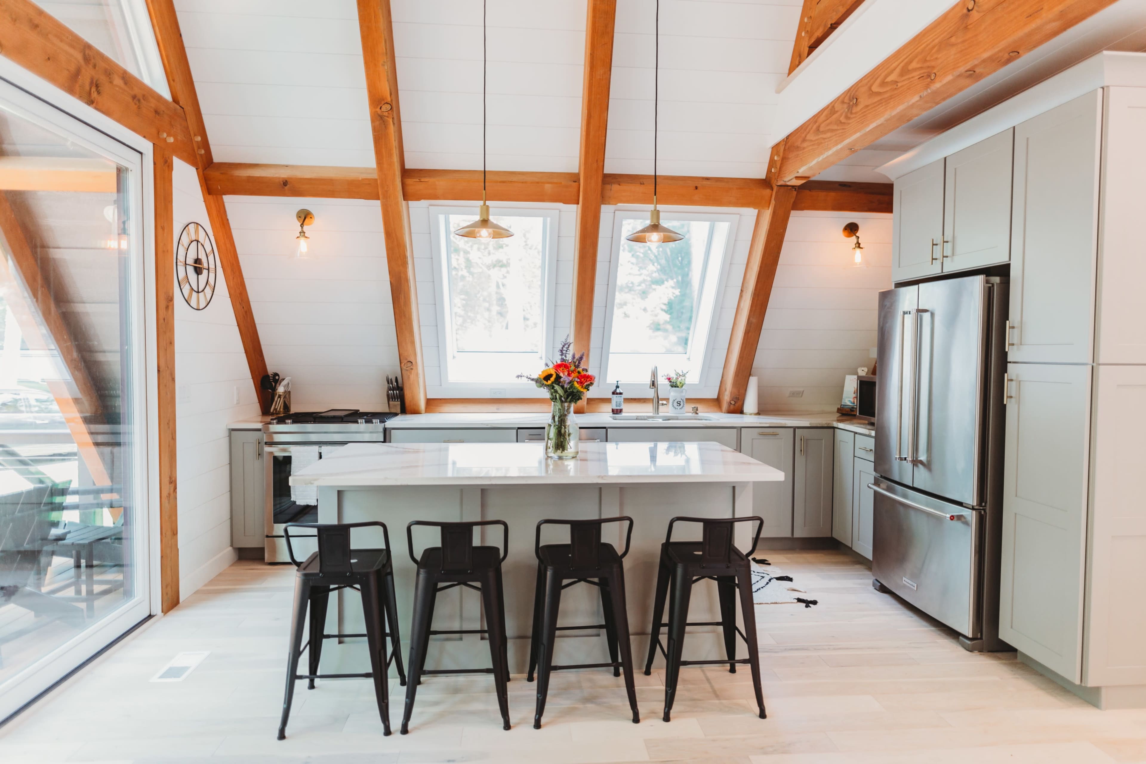 The image shows a modern kitchen featuring wooden beams, a central island with black bar stools, and large windows allowing natural light.