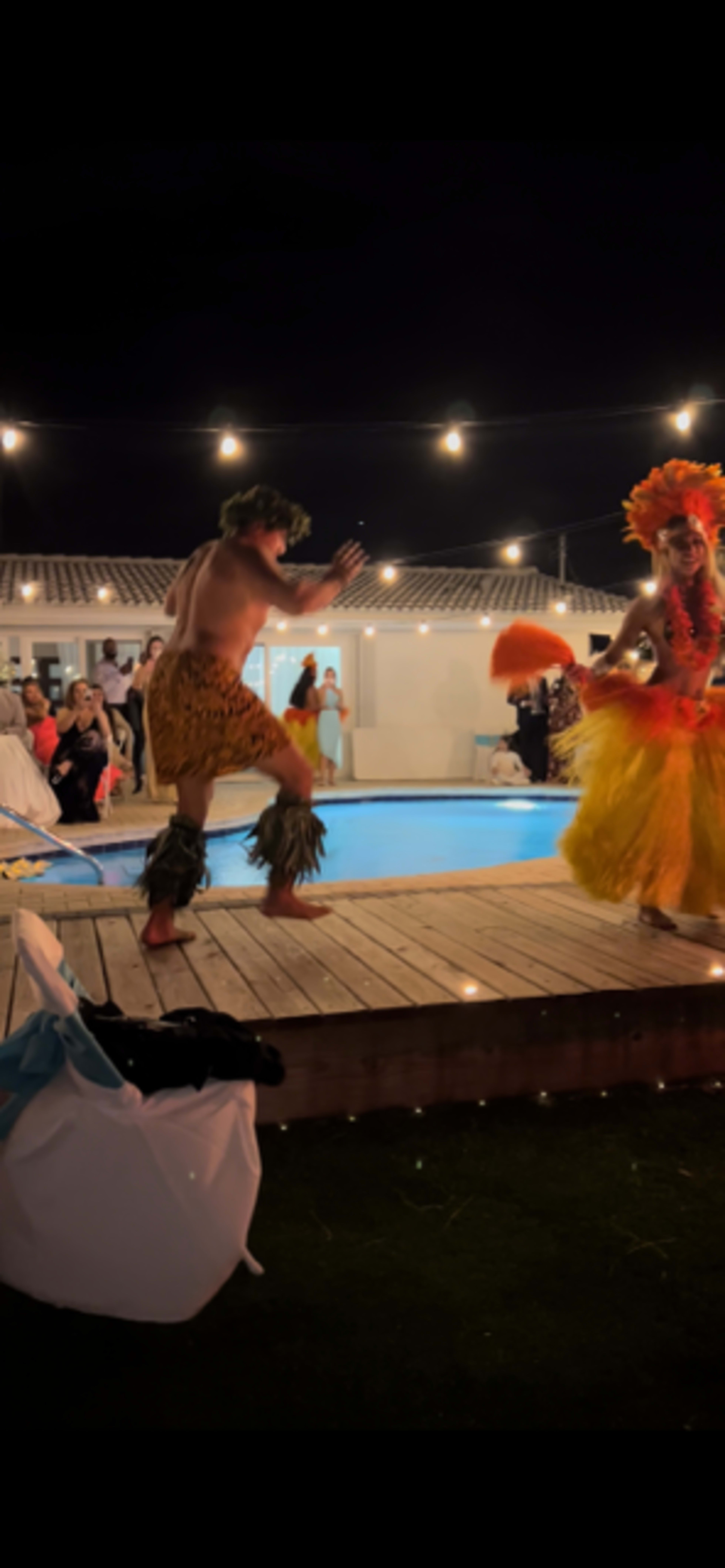 A performer in traditional attire dances alongside another performer near a pool illuminated by string lights at a nighttime event.