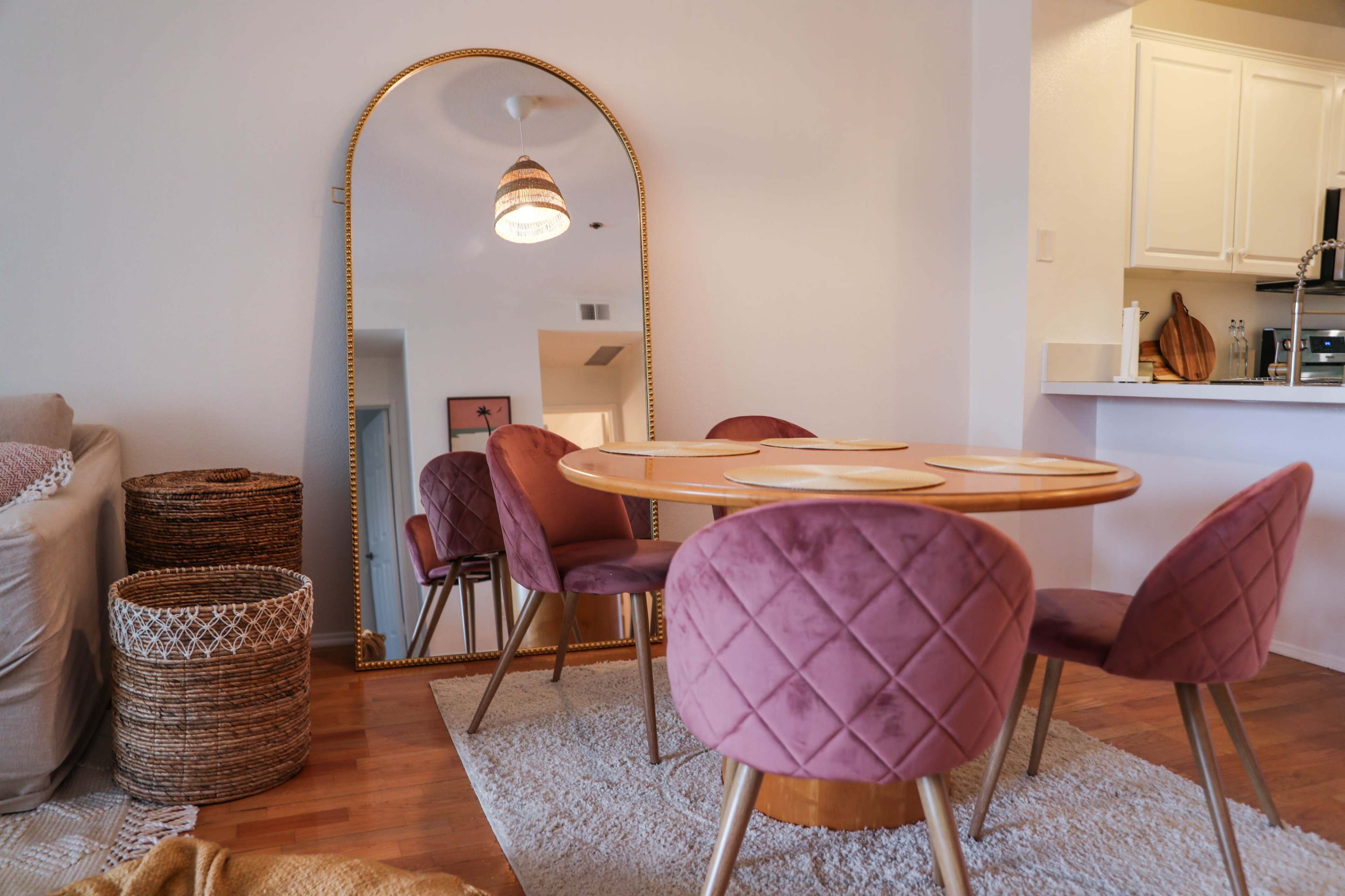 A dining area features a round table surrounded by pink upholstered chairs and a large mirror reflecting the space.