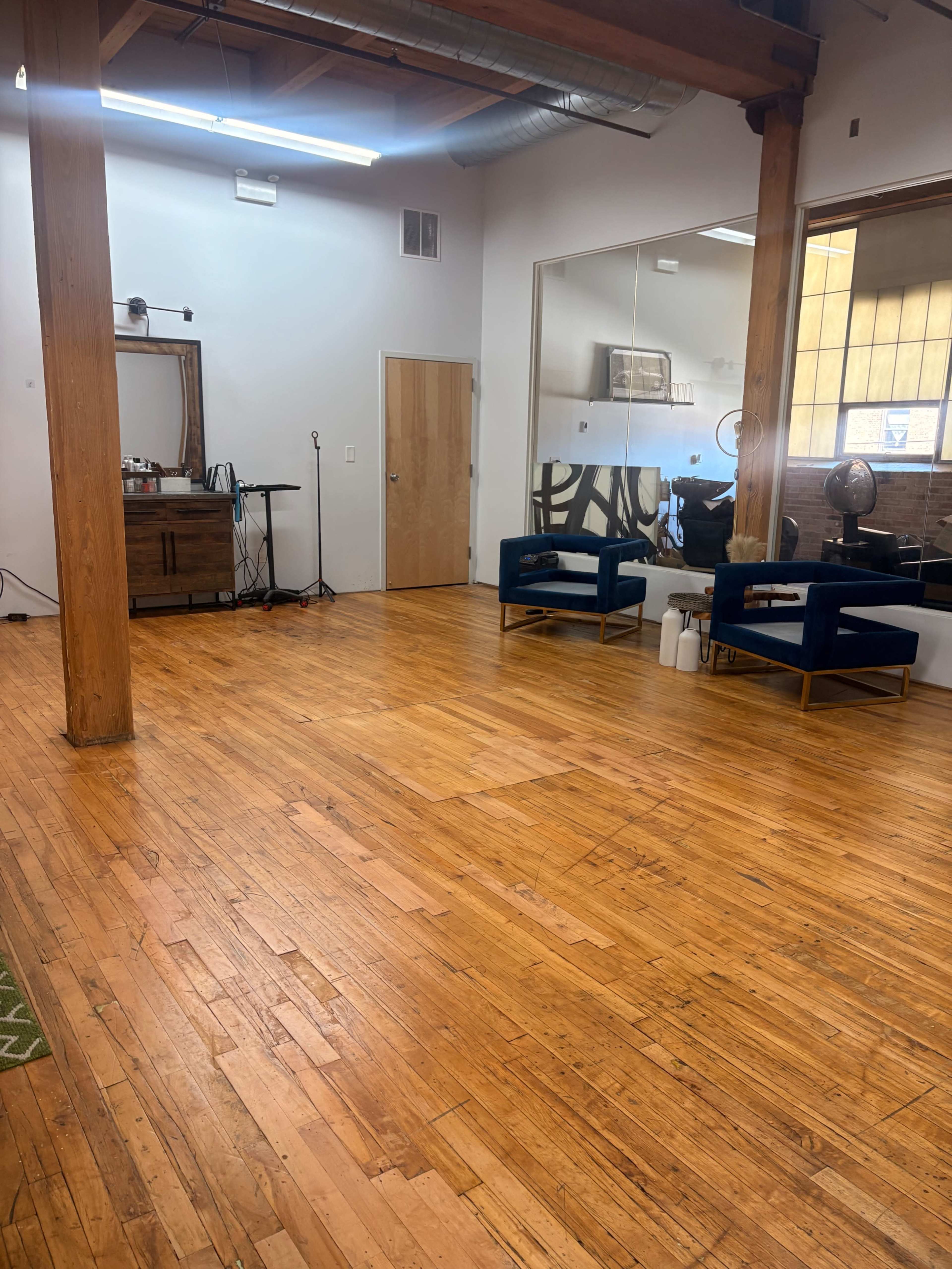 The image shows a spacious room with wooden flooring, featuring two blue chairs, a mirror on the wall, and a wooden cabinet.