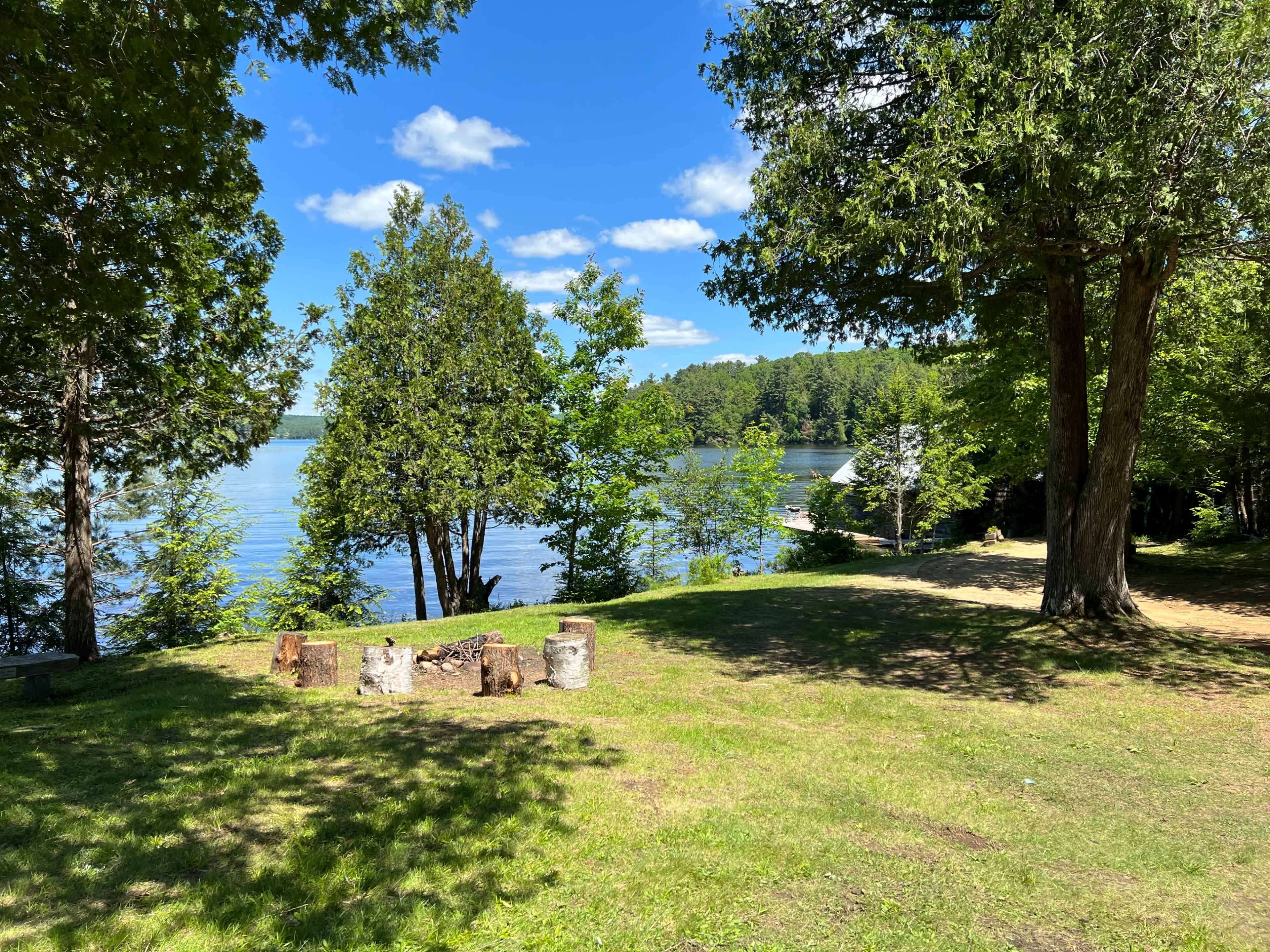 A clear lake is surrounded by green trees and a grassy area with a fire pit in the foreground.