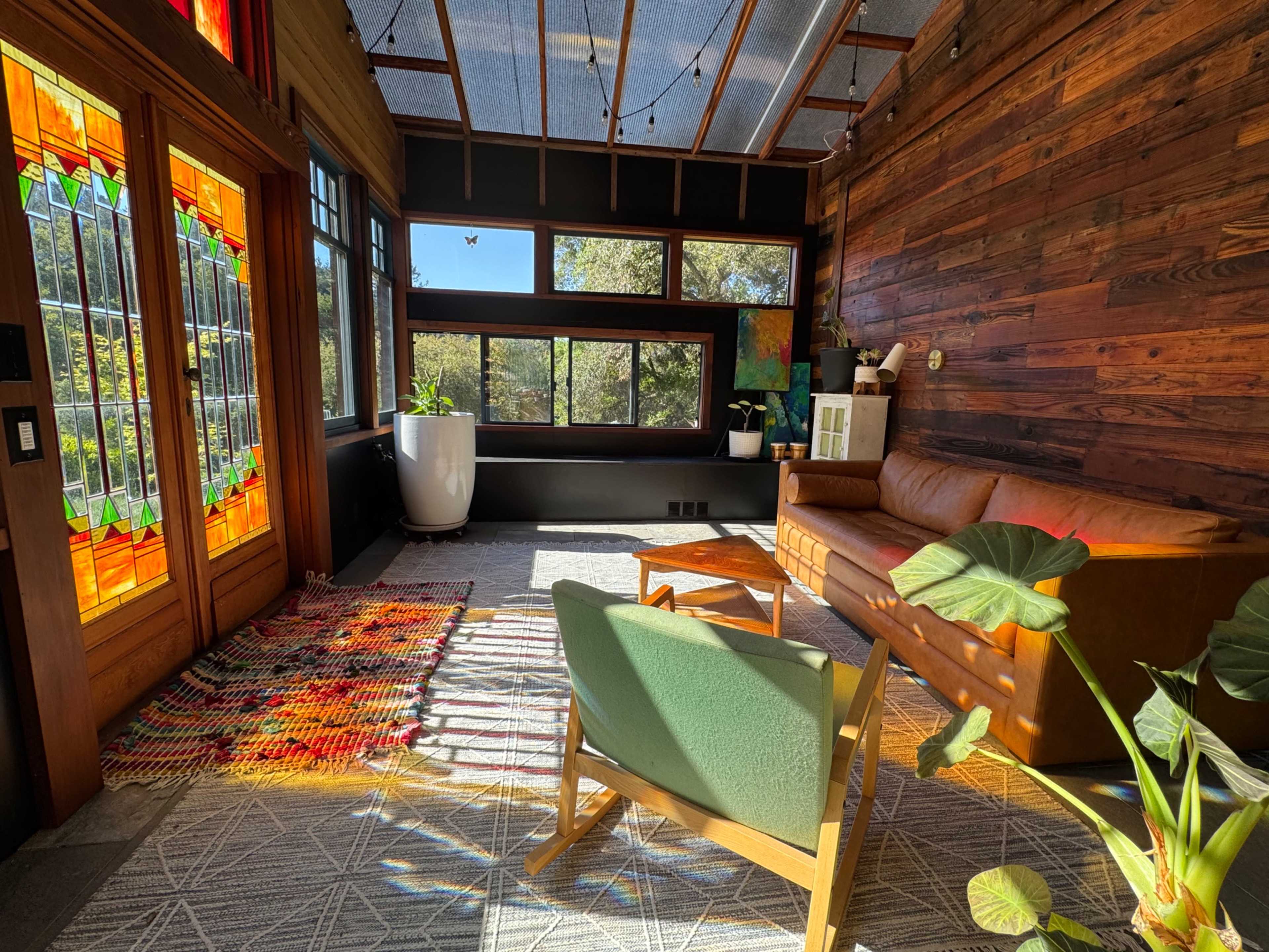 The image shows a cozy living space with a large stained glass door, wooden walls, a brown leather sofa, and plants inside.