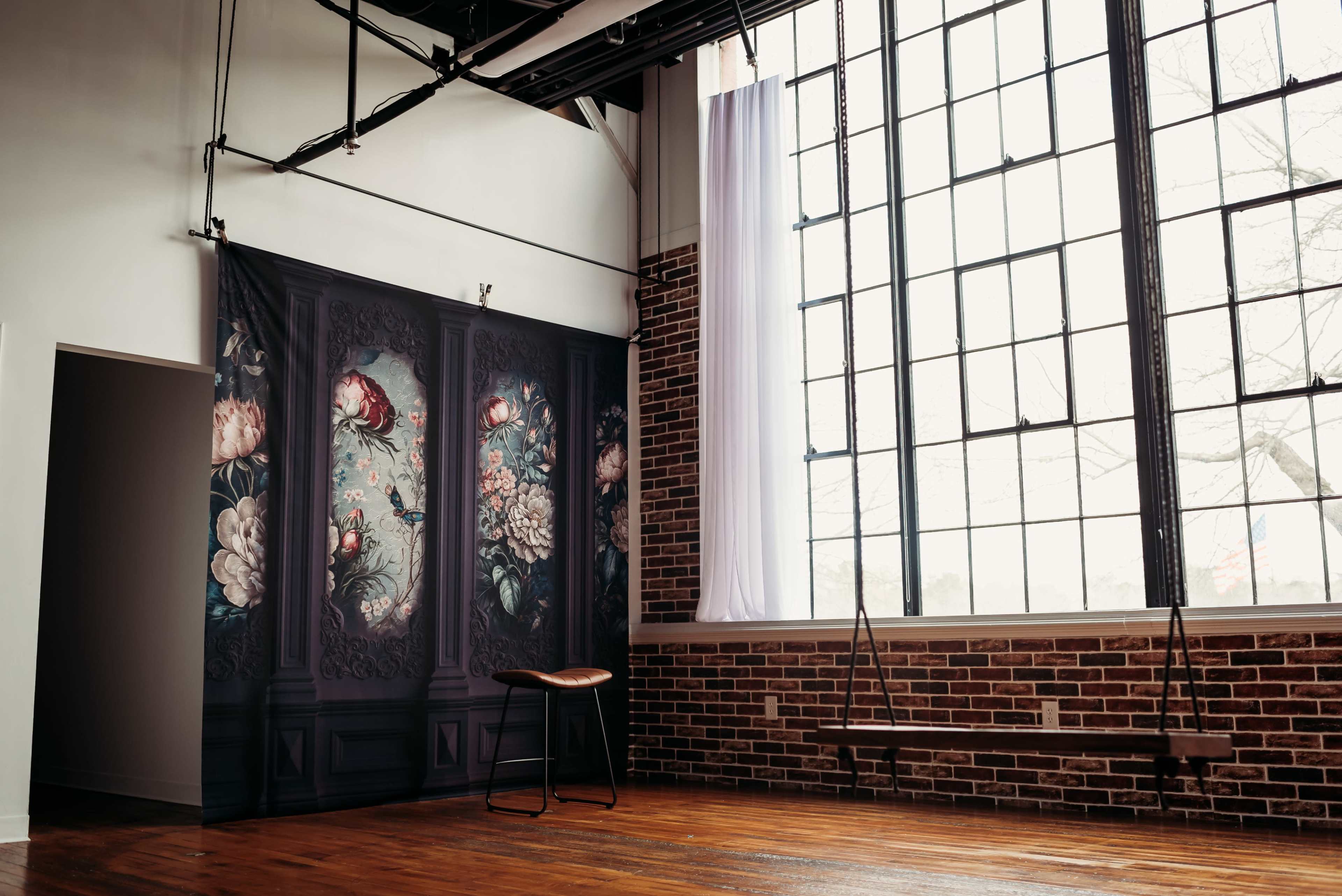 The image shows a spacious room with exposed brick walls, large windows allowing natural light to filter in, and a decorative mural on one wall beside a wooden stool.