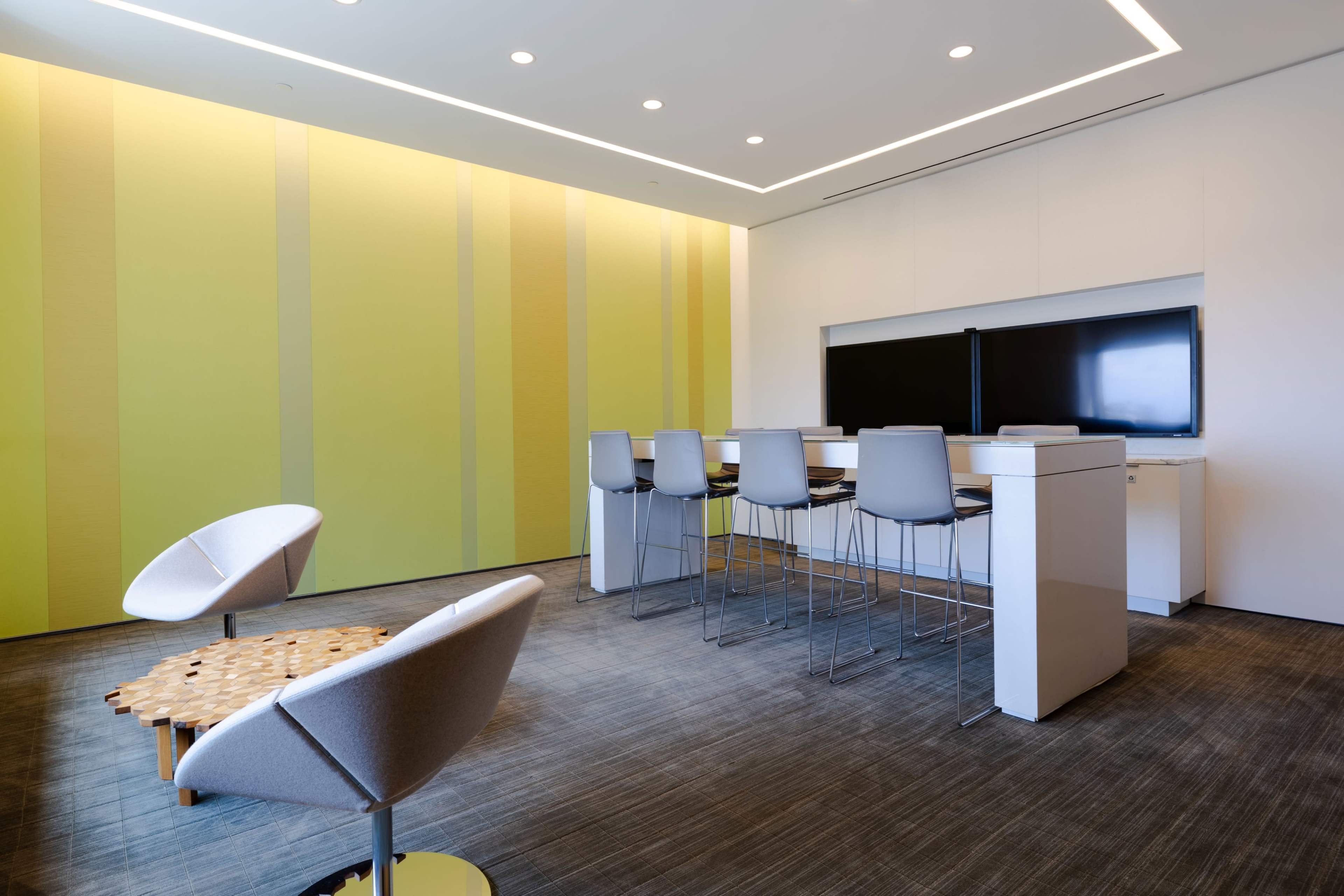 The image shows a modern meeting room with yellow-striped walls, high tables, and stools, along with a couple of contemporary chairs.