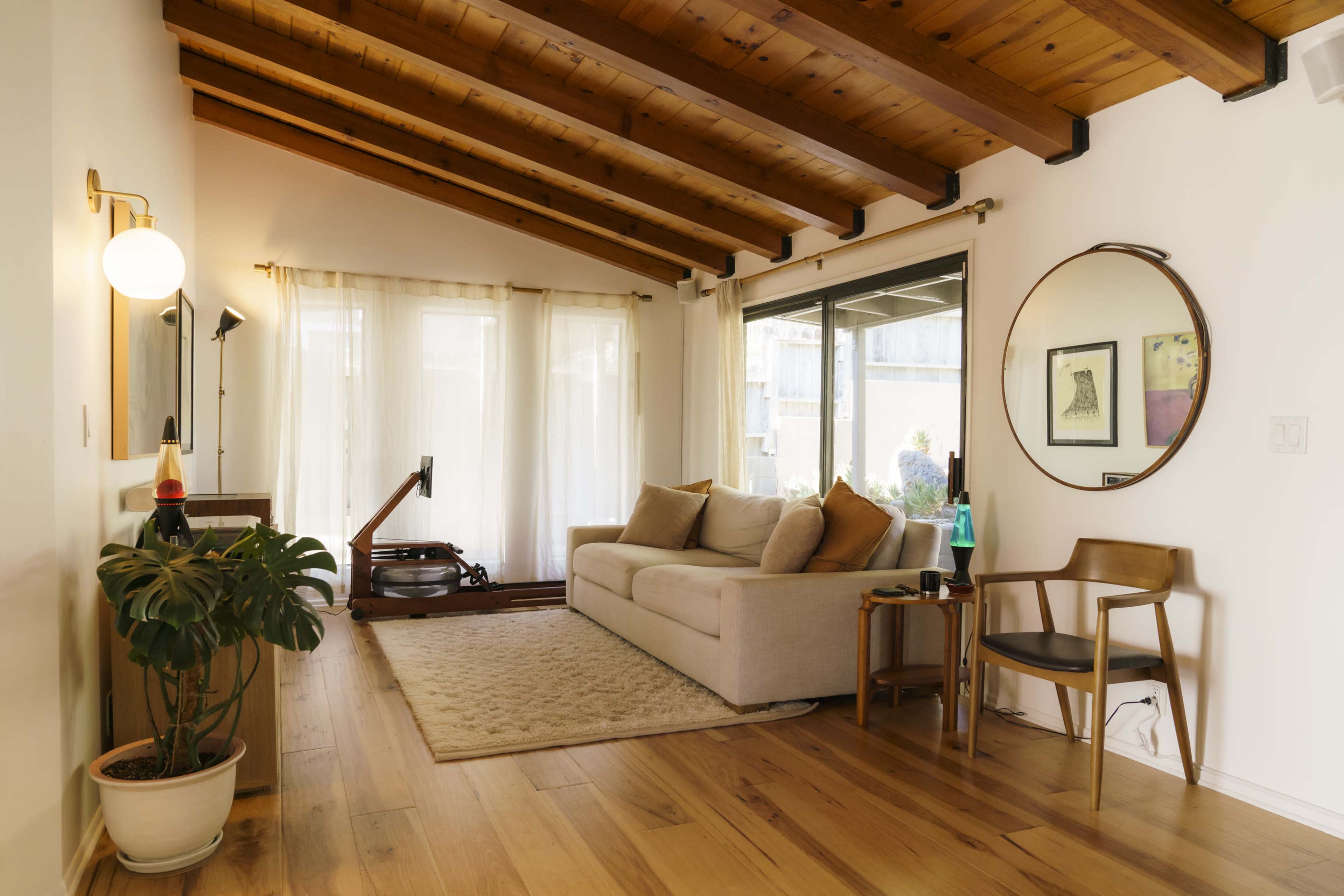 A living room with a light-colored sofa, large windows allowing natural light, wooden ceiling beams, and a small plant beside a mid-century modern chair.