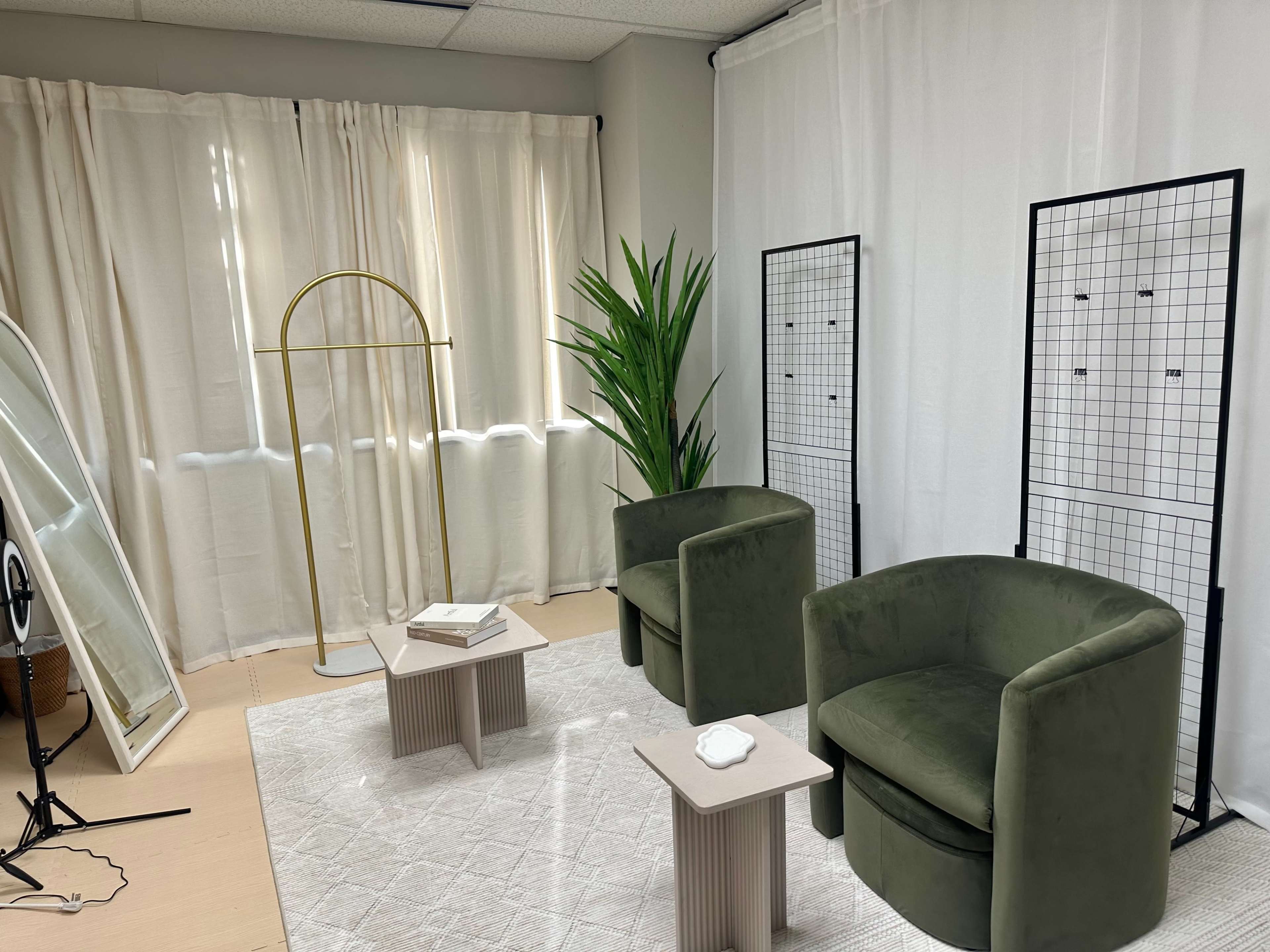 The image shows a small interior room with two green chairs, a mirror, a plant, and two grid-like display stands against a white backdrop.