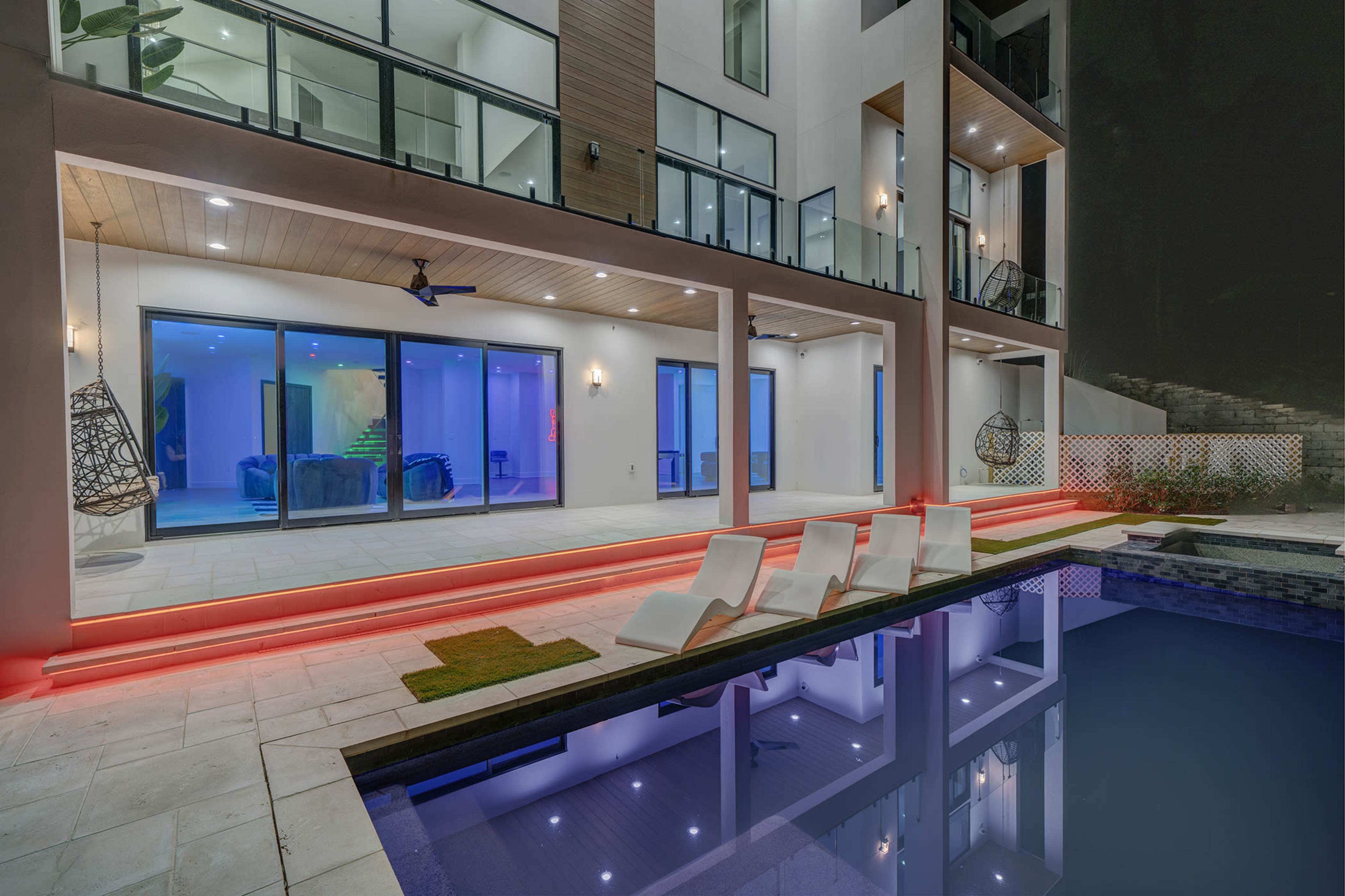 The image shows a modern two-story house with large glass windows, an outdoor pool, and white lounge chairs, illuminated by red lighting along the patio.