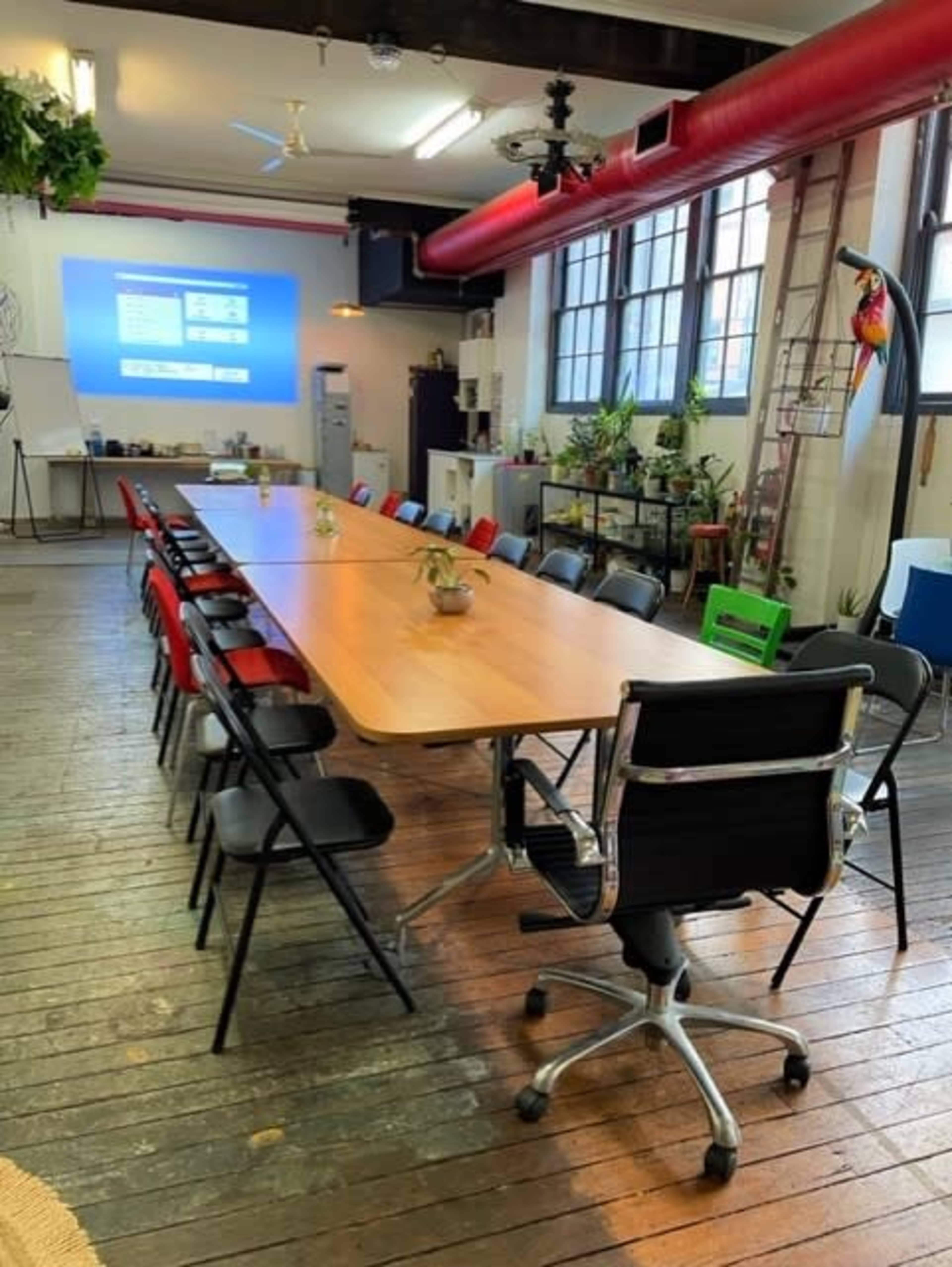 A long wooden table surrounded by various chairs is set up in a well-lit room with a projector screen and greenery in the background.