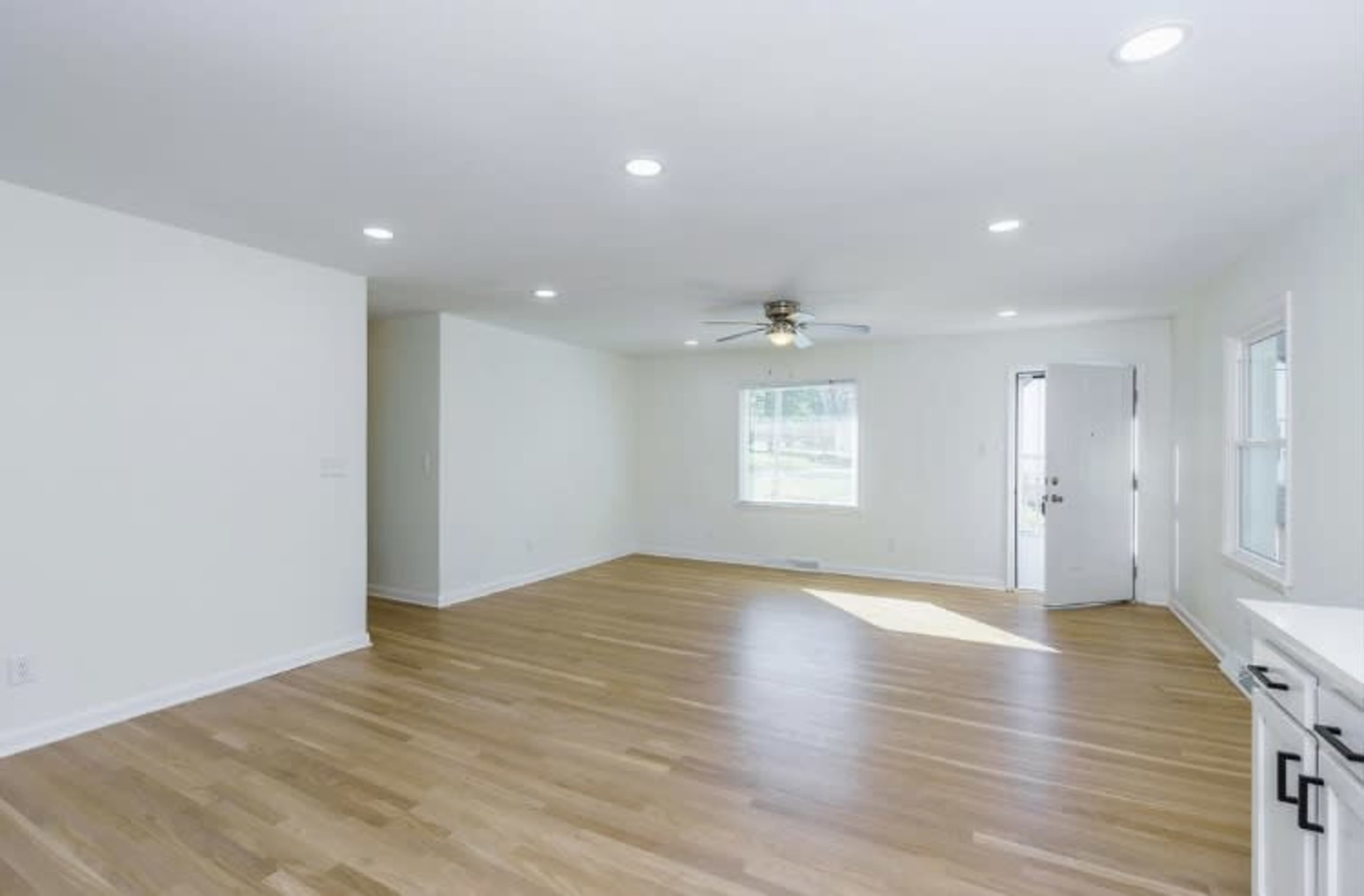 A spacious, empty living room with light-colored walls and wooden flooring, featuring a ceiling fan and a window next to a door.