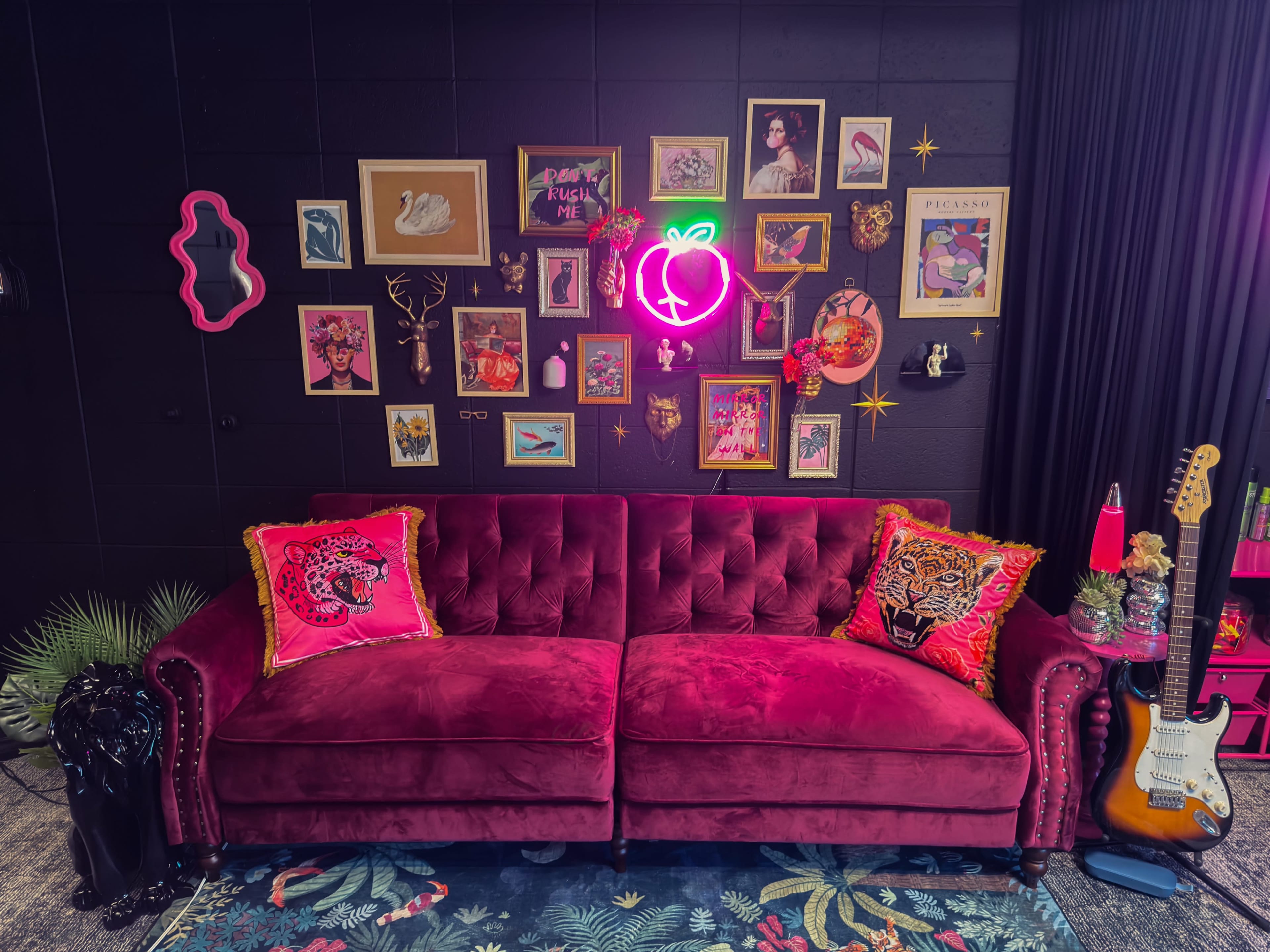 A vibrant room features a plush pink sofa adorned with decorative pillows, against a wall decorated with framed artwork and a neon peach sign.