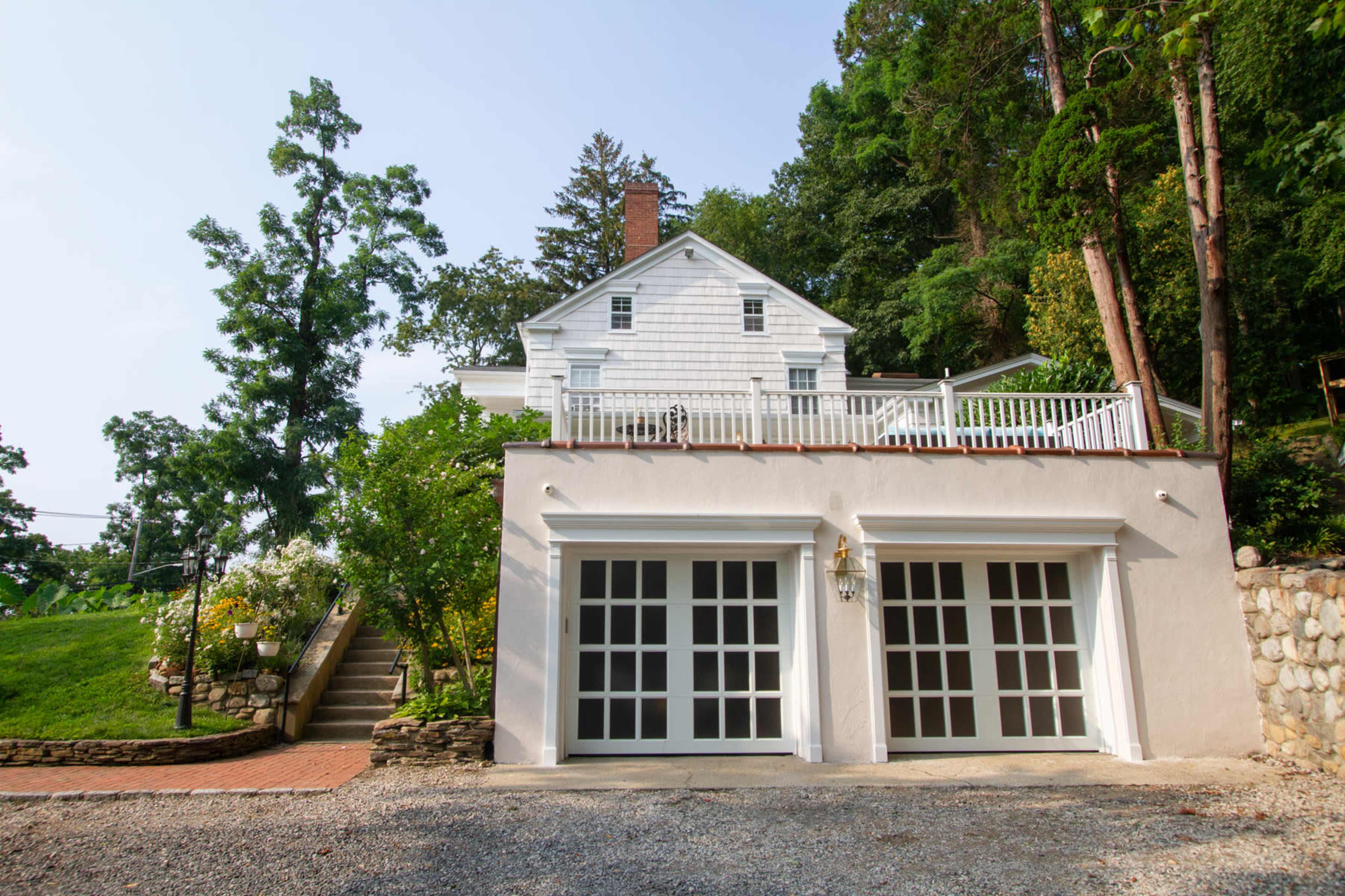 A two-story white house with a porch is situated on a hillside, featuring a stone pathway leading to it and a two-car garage at the front.