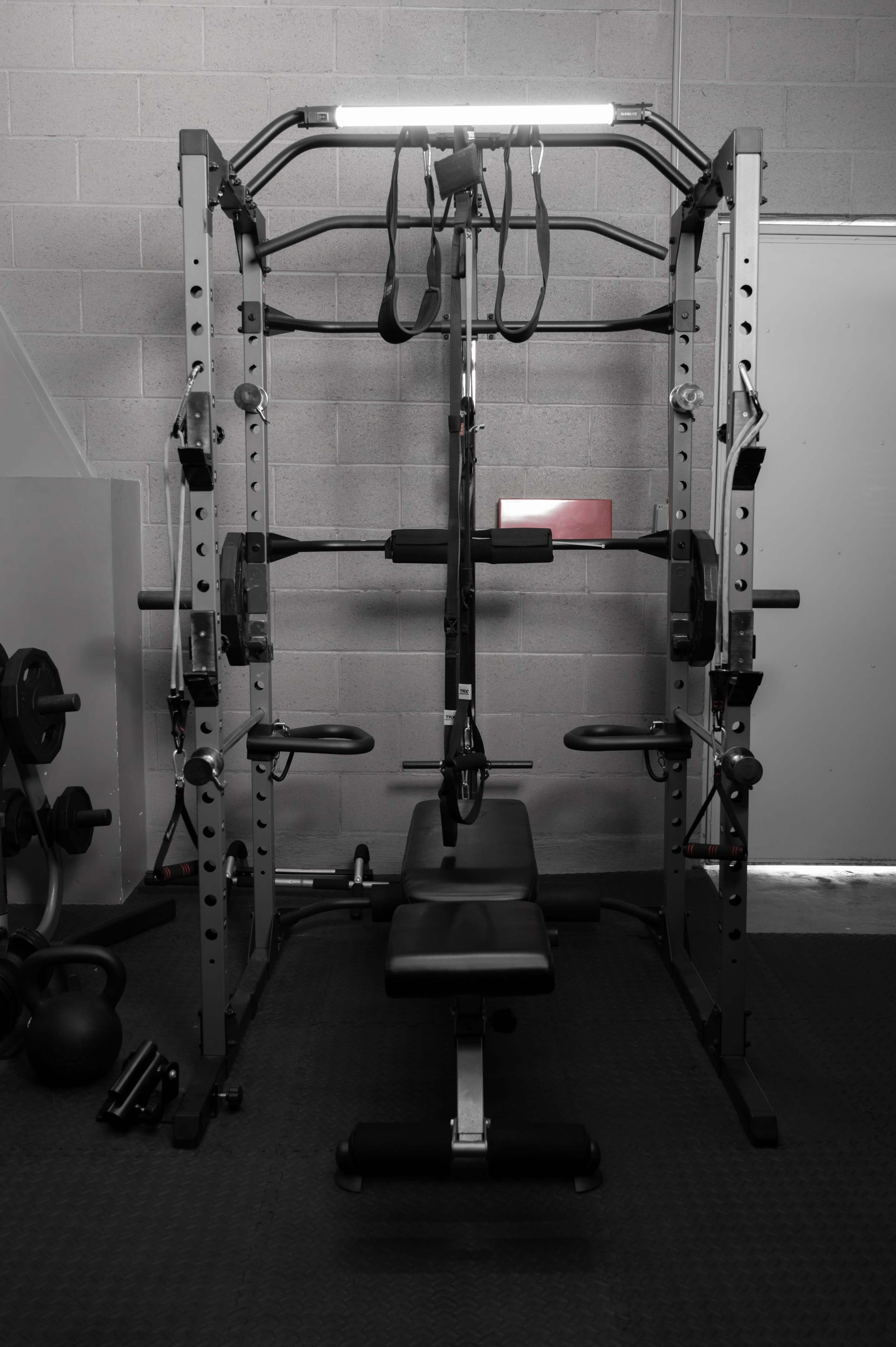 A gym equipment setup consisting of a weight rack with a bench and various attachments against a gray wall.