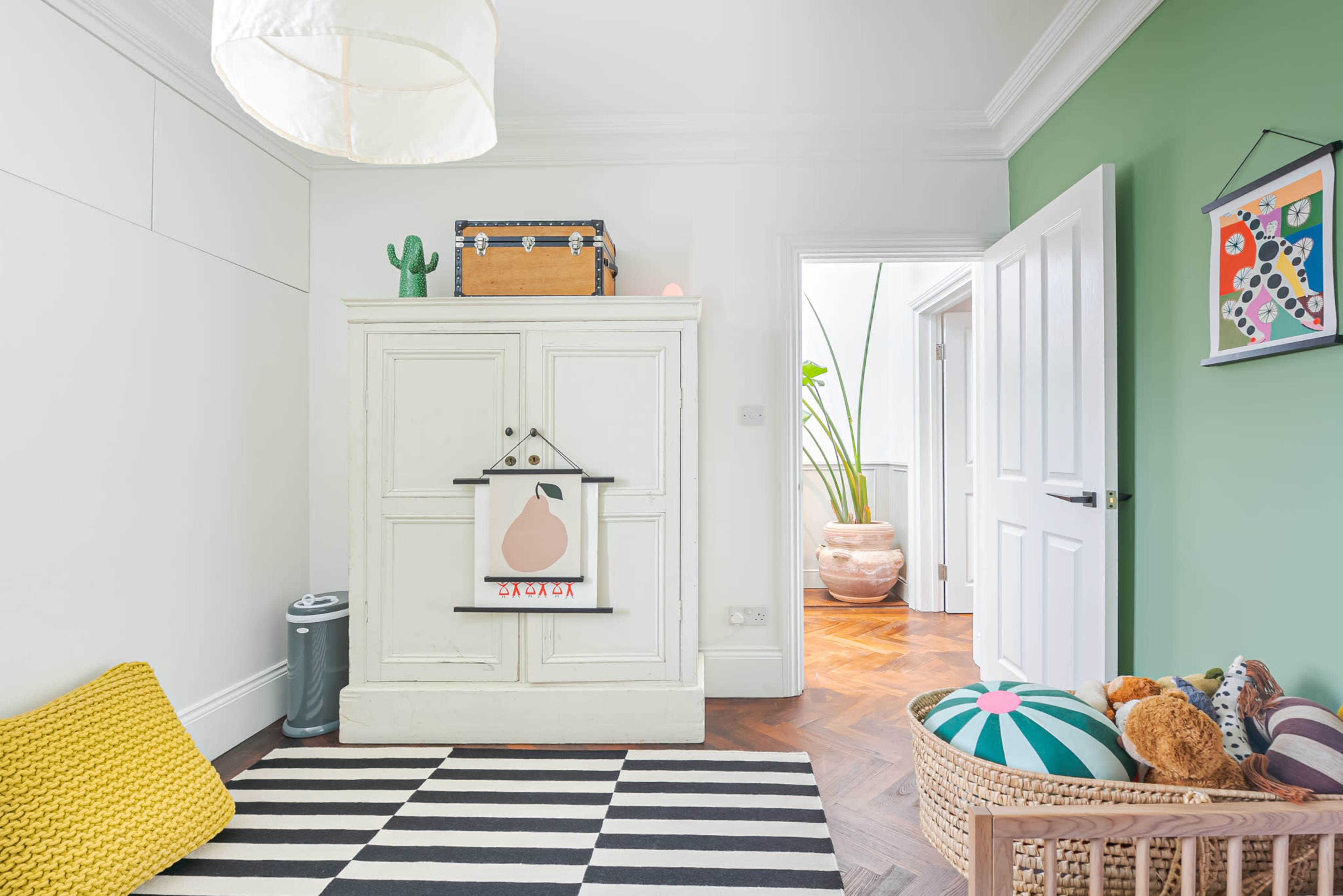 The image shows a room featuring a green wall, a wardrobe with a hanging print, a striped rug, and a basket filled with plush toys.