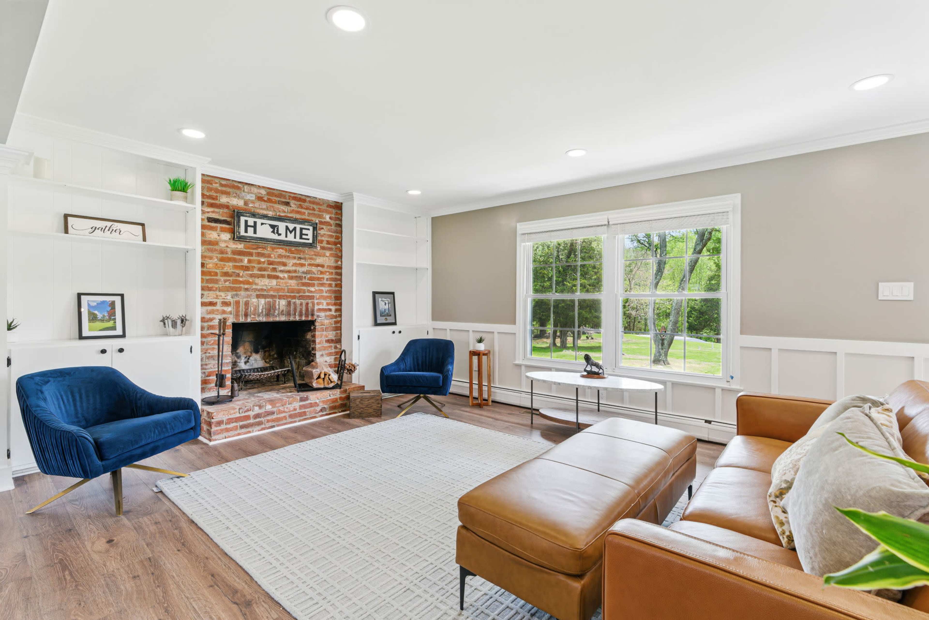 The living room features a brick fireplace, two navy blue chairs, and a brown leather sectional sofa, with large windows allowing natural light to fill the space.