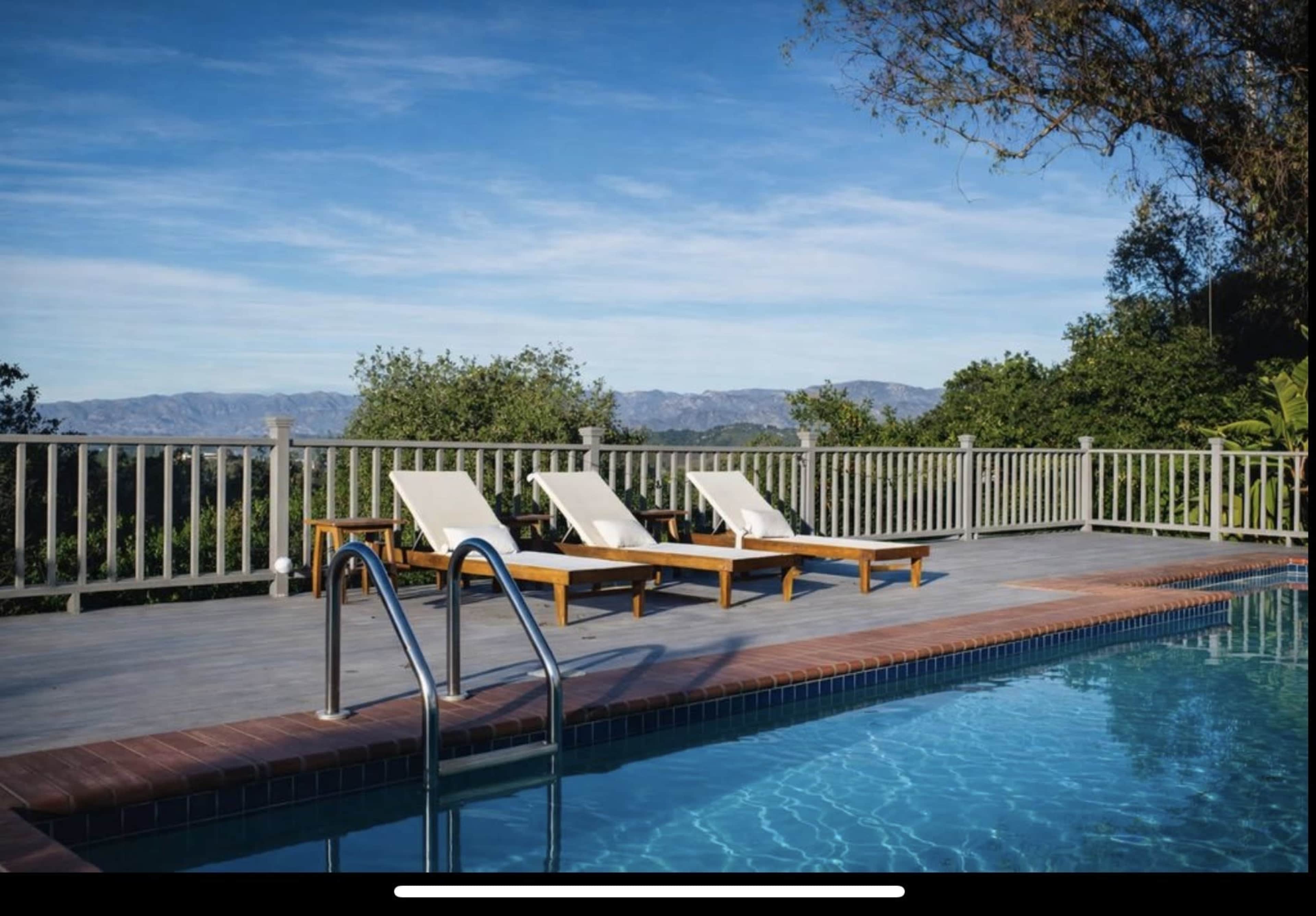 A serene outdoor scene features a swimming pool with a ladder, flanked by two lounge chairs on a wooden deck overlooking a mountainous landscape.