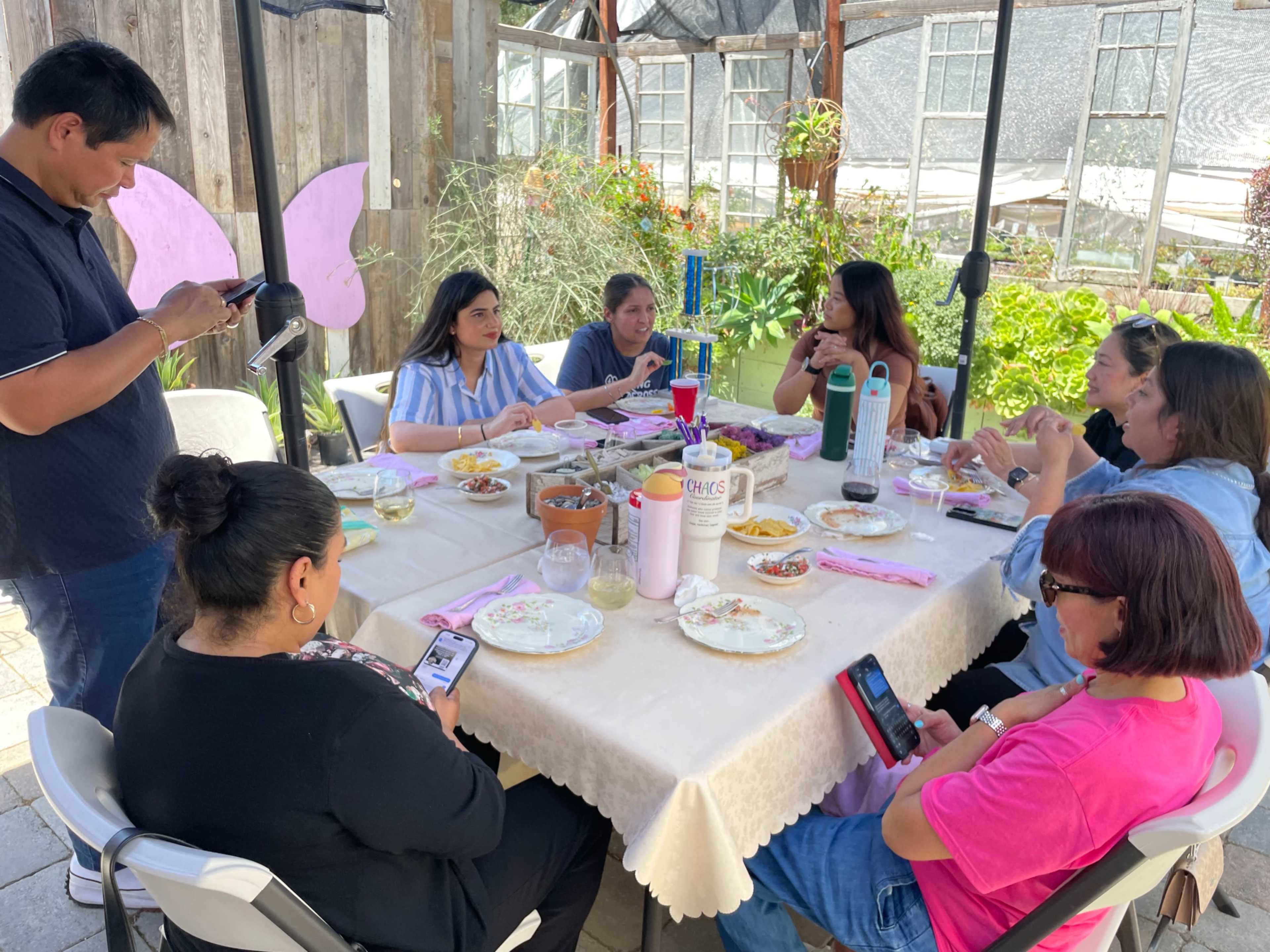 A group of eight people gathers around a table with plates of food and drinks in an outdoor setting.