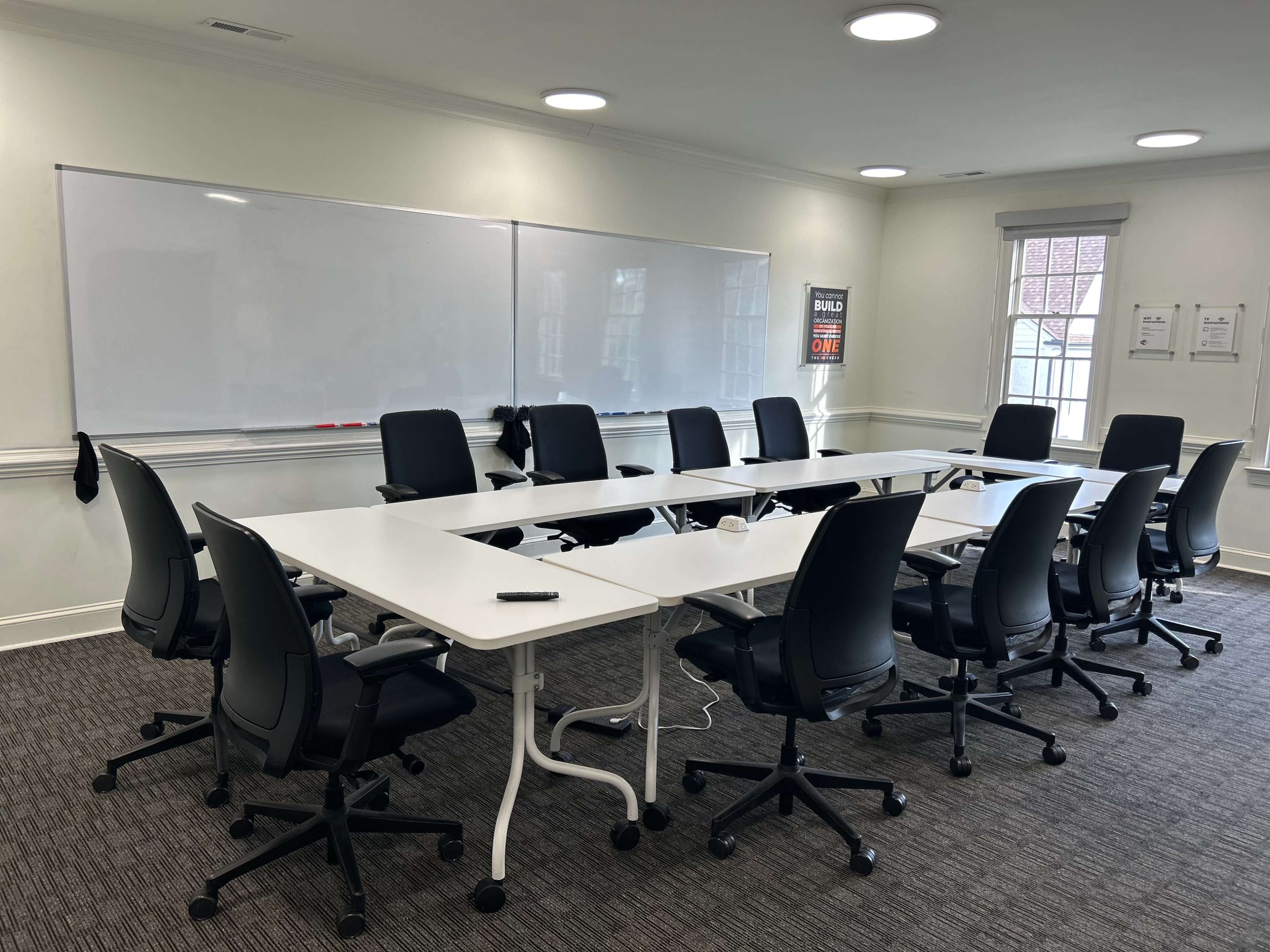 A conference room features a U-shaped arrangement of white tables surrounded by black office chairs, with whiteboards on the walls and natural light coming from windows.