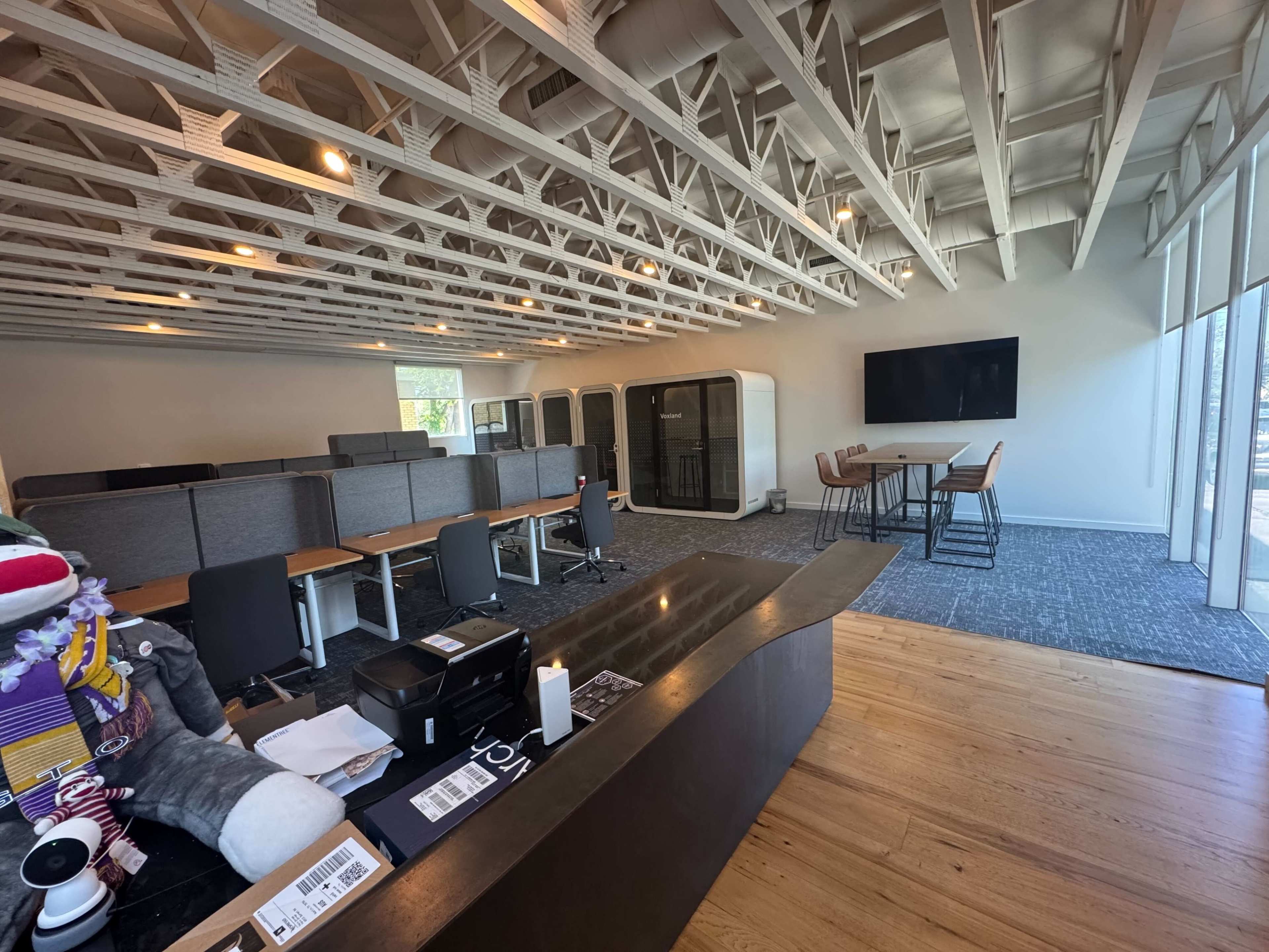 The image shows a modern office space with several cubicles, a collaborative table, and a large screen mounted on the wall.