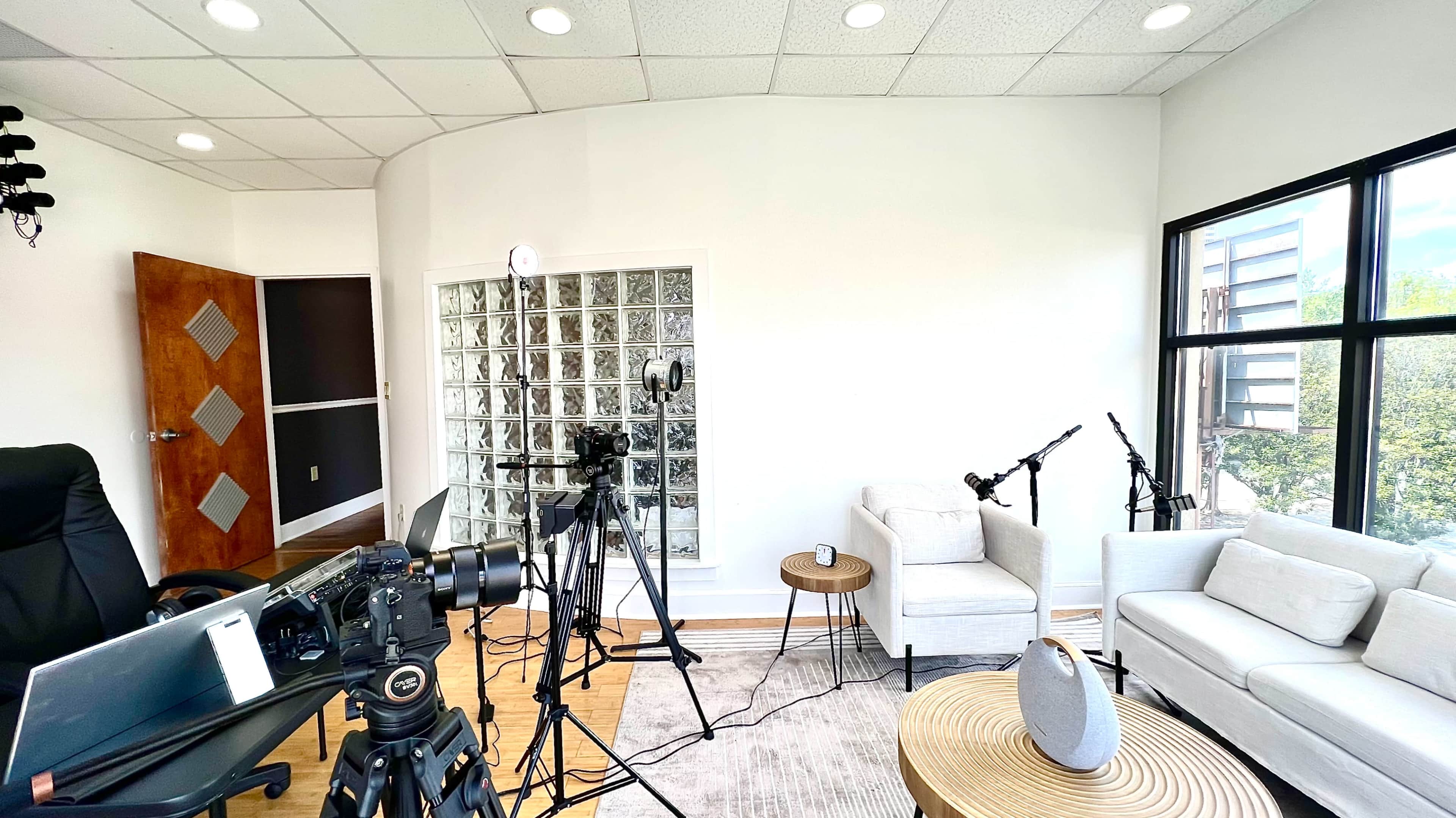 A modern, minimalist room equipped with multiple cameras and audio equipment, featuring a light-colored sofa and a glass block wall.