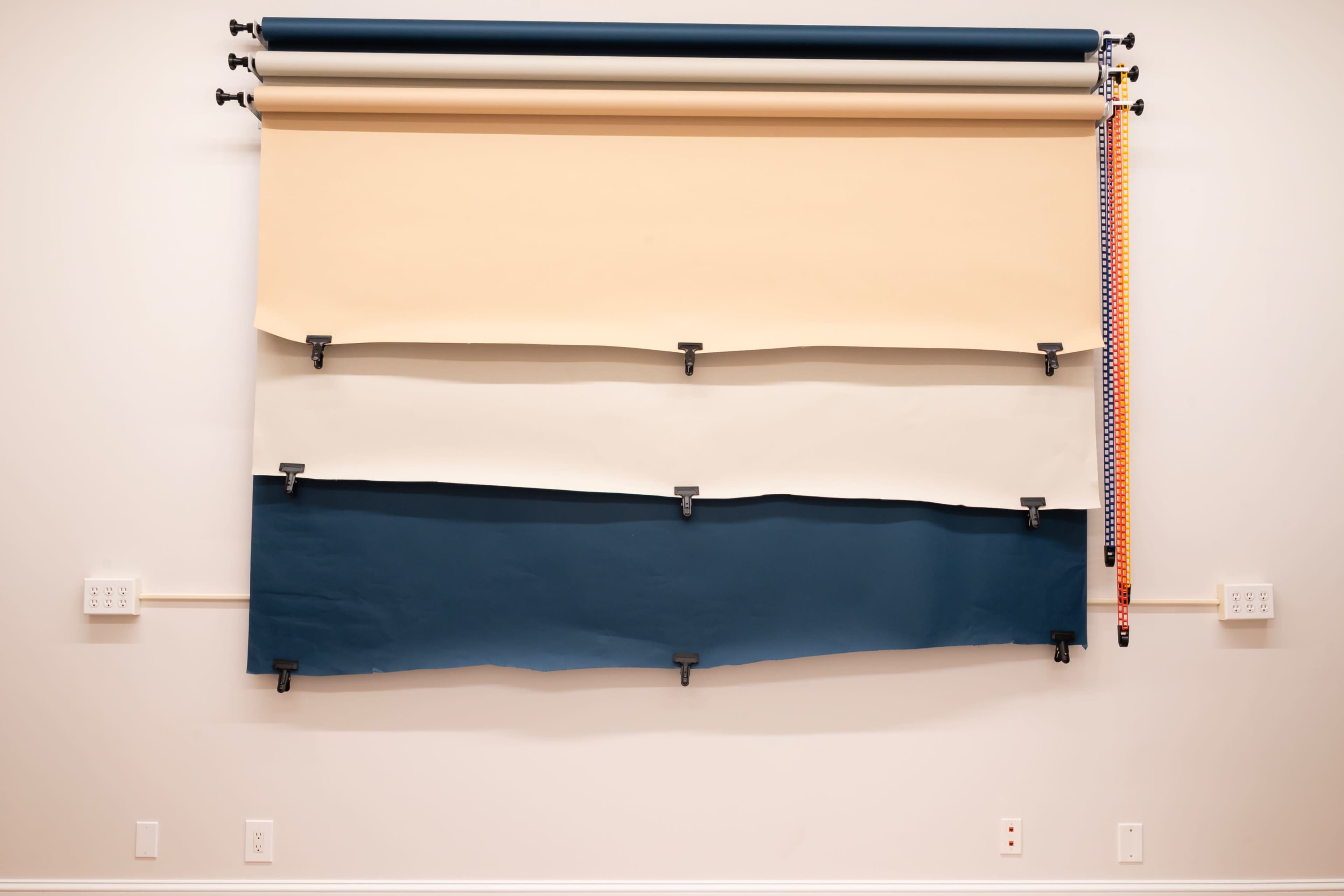 The image features three rolls of colored paper—beige, cream, and dark blue—mounted on a wall with brackets and a patterned strap hanging beside them.