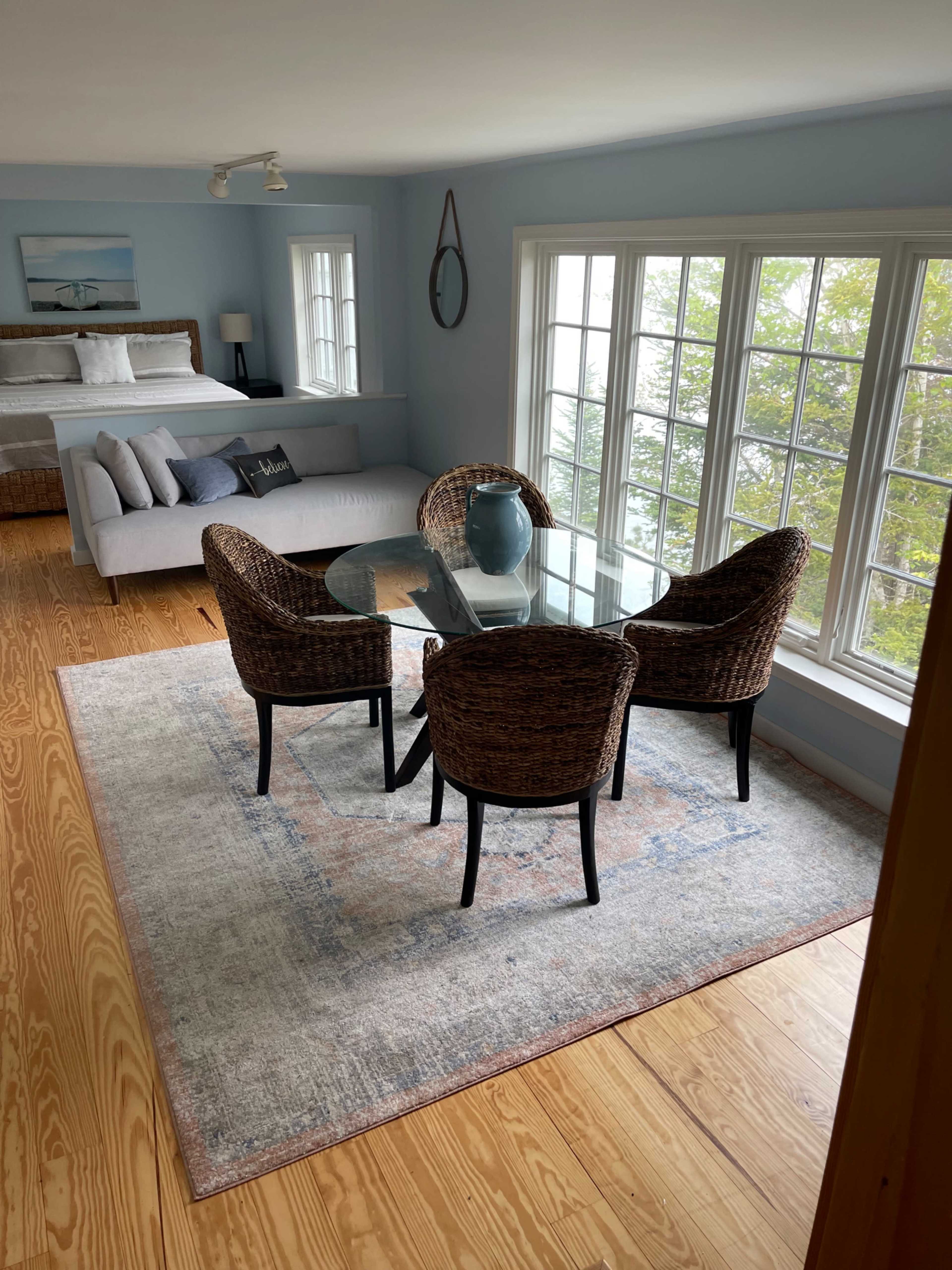 A cozy indoor seating area with a glass-top table surrounded by four wicker chairs, positioned near large windows that offer natural light.