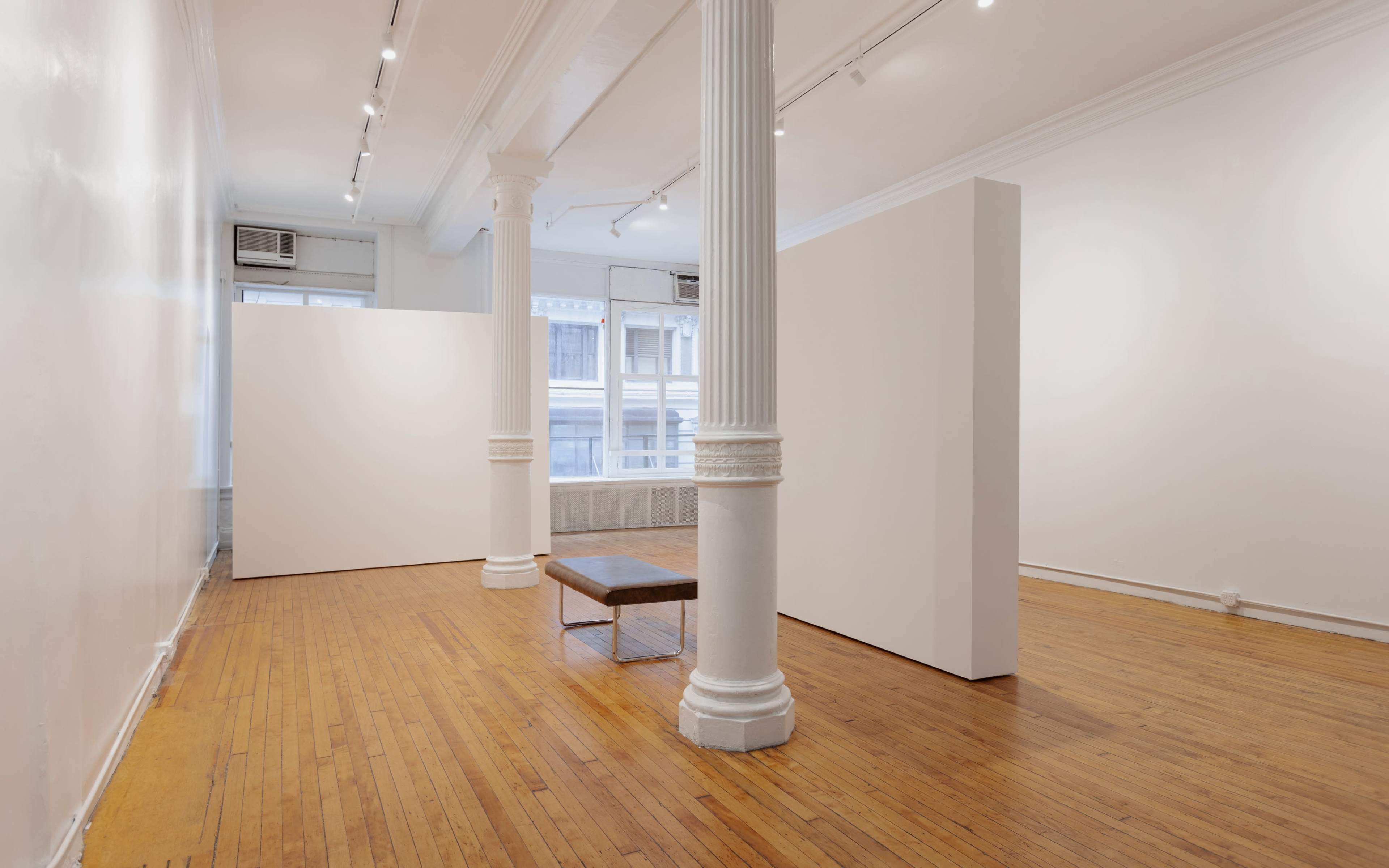The image shows a minimalist art gallery with white walls, wooden flooring, and a simple bench in front of a partitioned space.