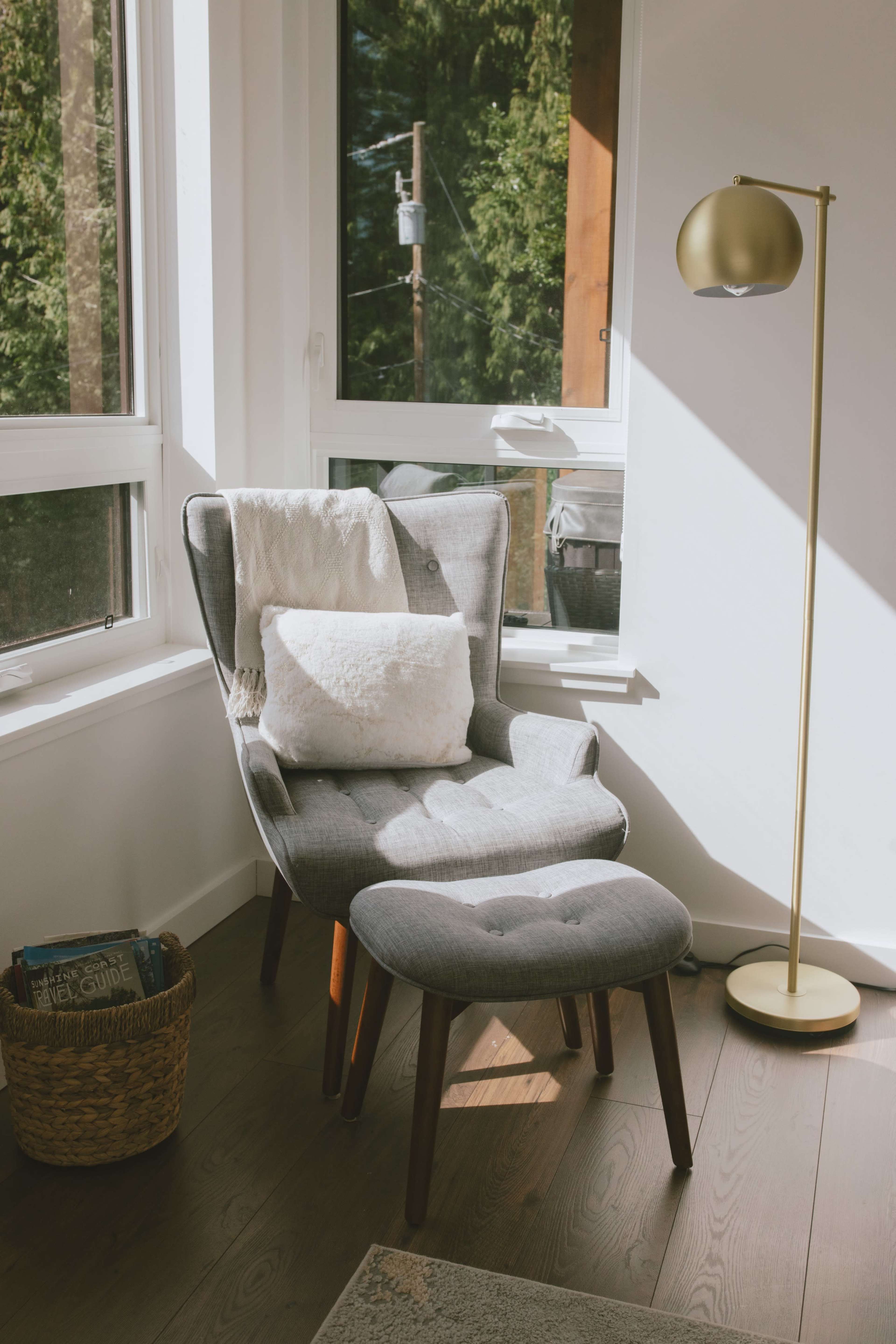 A modern gray armchair with a matching ottoman sits beside a floor lamp in a well-lit corner with large windows and a woven basket nearby.