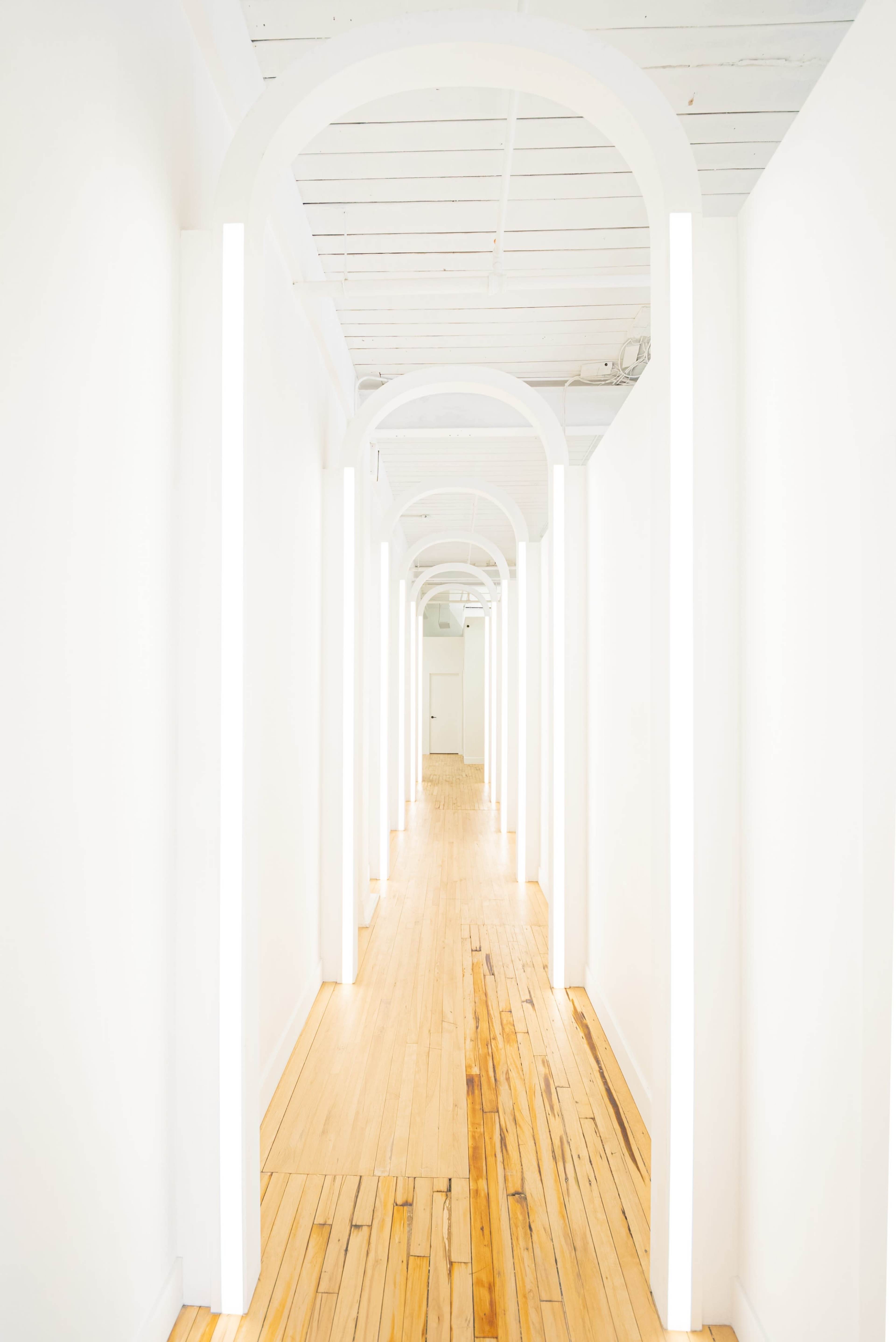 A long, narrow hallway lined with arches and illuminated by vertical light fixtures against white walls and a wooden floor.