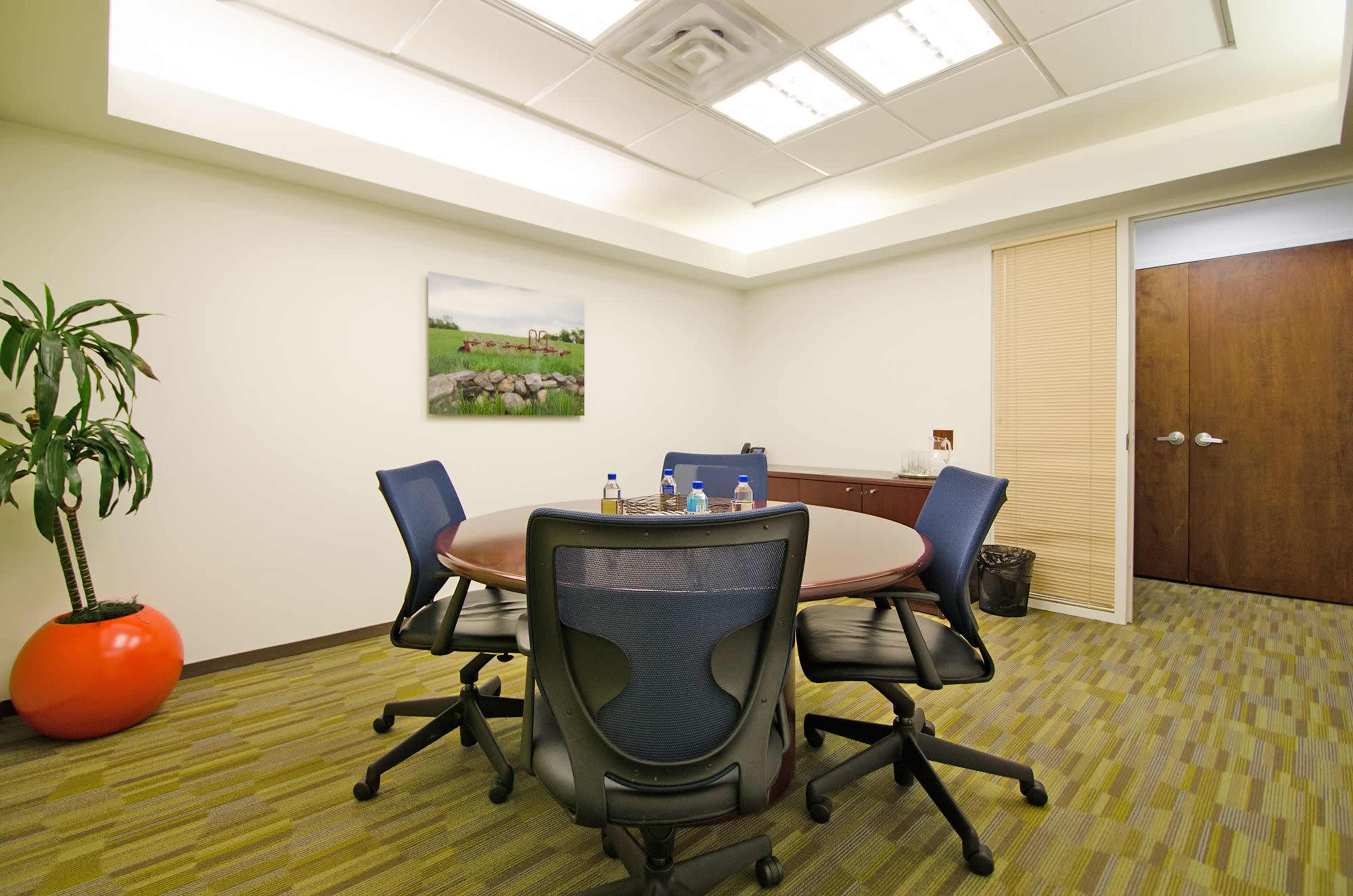 A conference room features a round table with four chairs, a plant, and a piece of artwork on the wall.