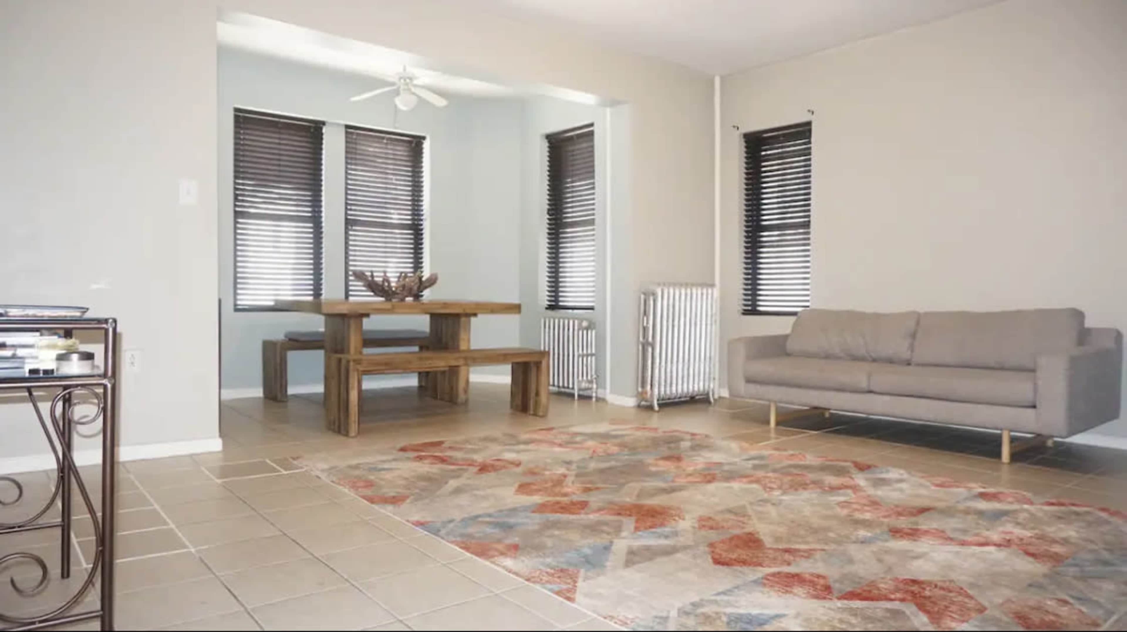 A spacious room featuring a wooden dining table, a gray sofa, a patterned area rug, and large windows with dark blinds.