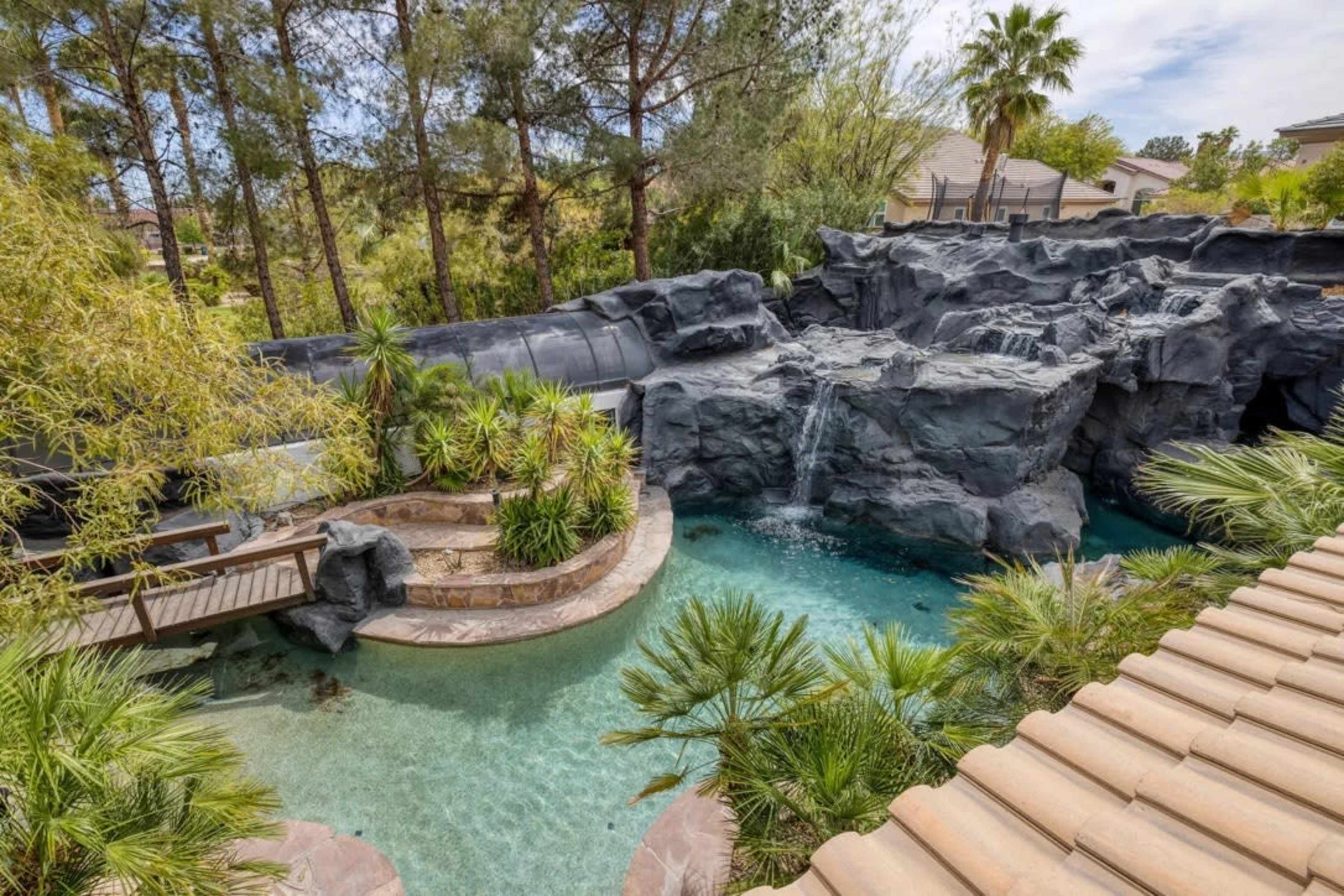 The image features a landscaped backyard with a pool that includes a rock formation and a waterfall, surrounded by palm trees and lush greenery.