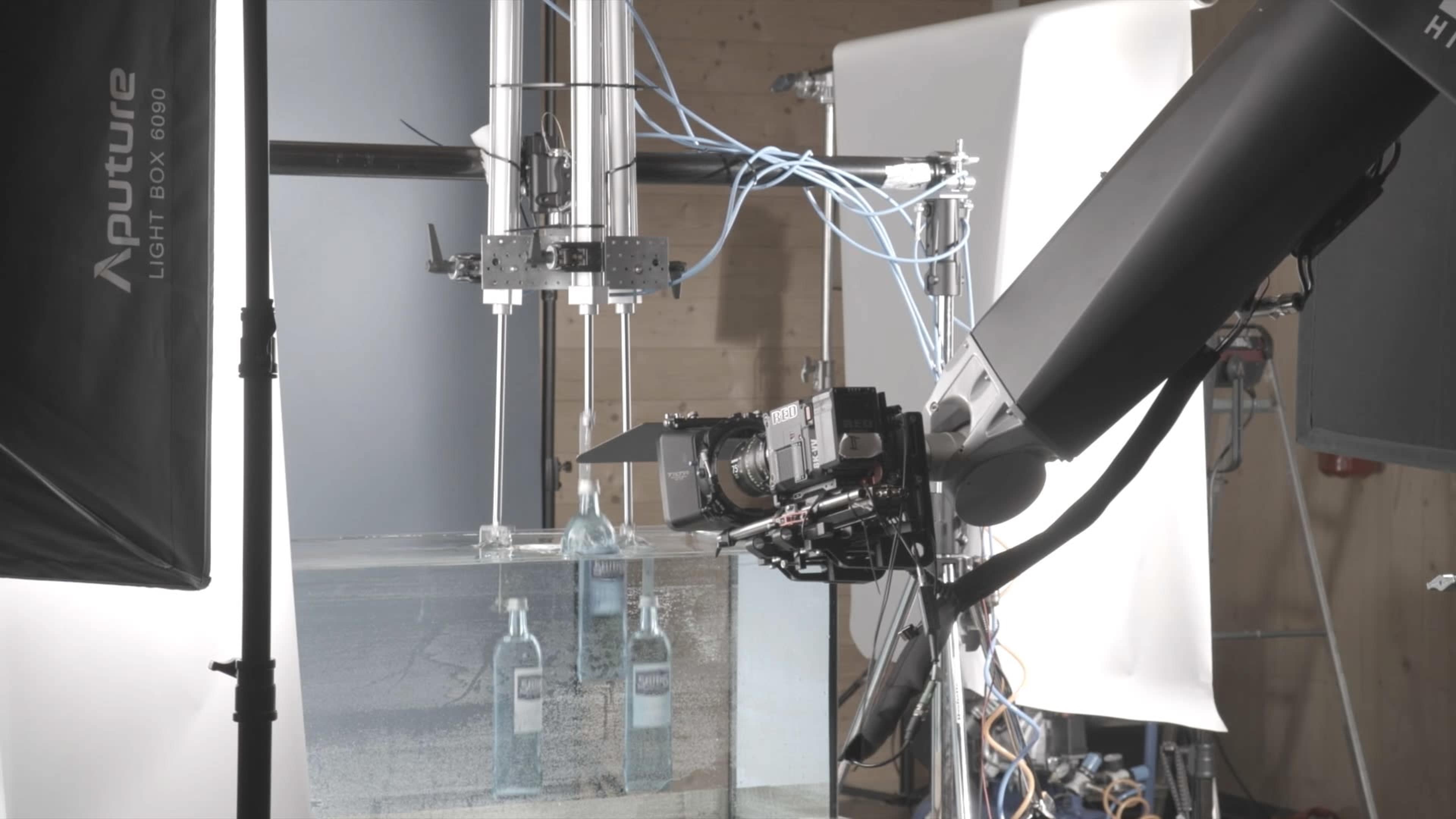 The image shows a filming setup with a robotic arm positioned over a tank containing bottles, alongside studio lighting equipment.