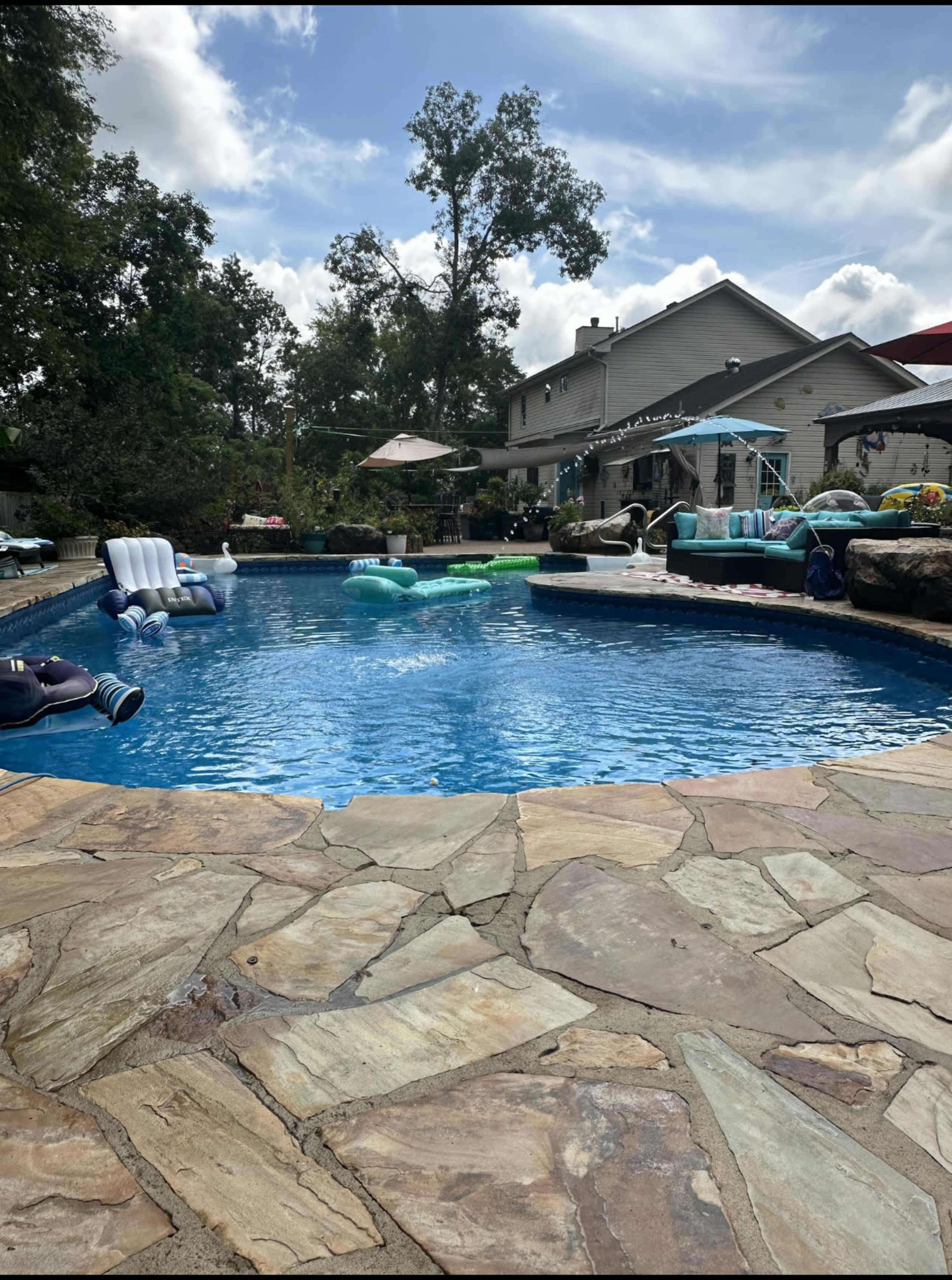 The image shows a residential backyard with a swimming pool surrounded by stone pavement and various floating devices.