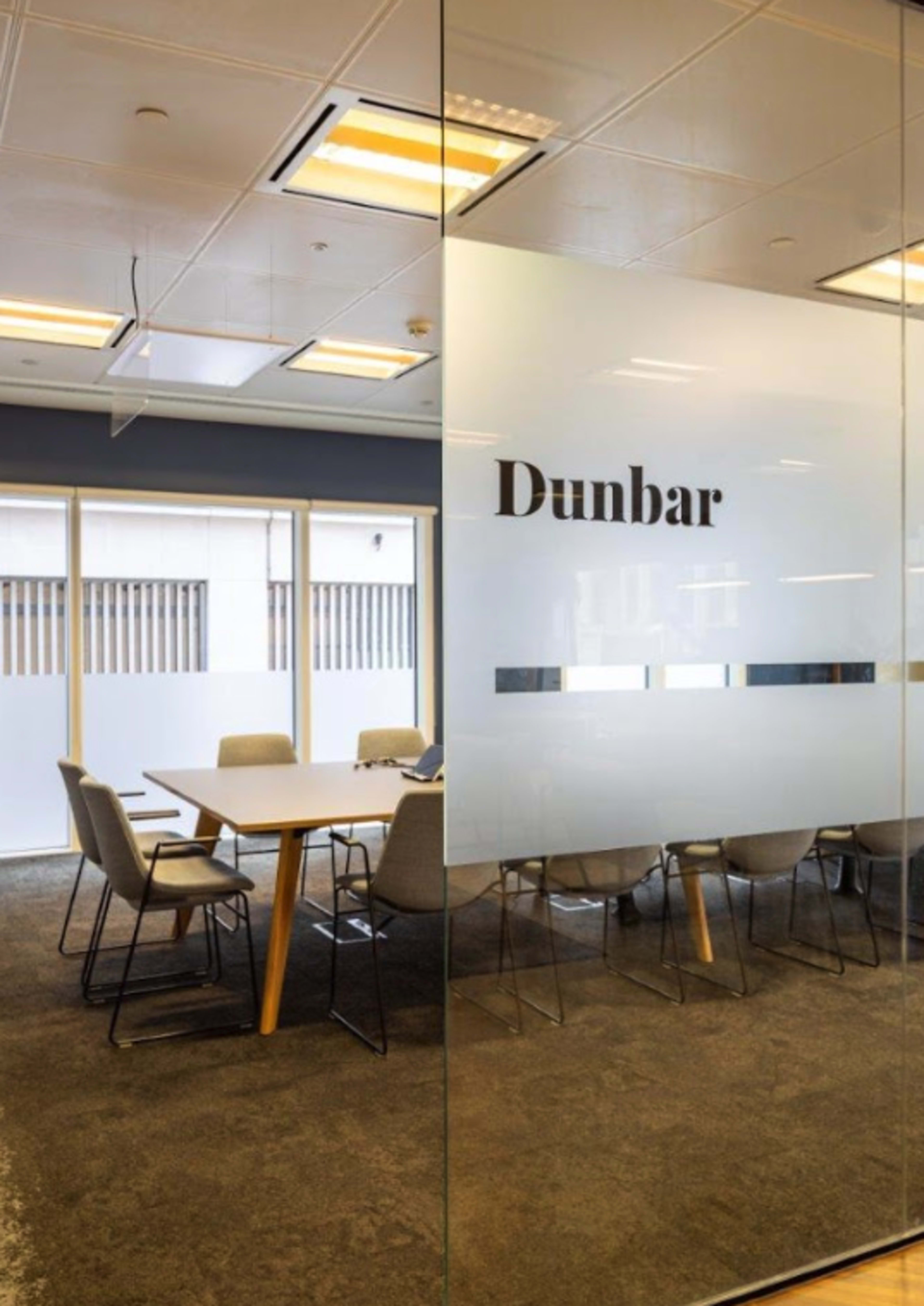 A glass-walled conference room labeled "Dunbar" with a large table surrounded by chairs and windows allowing natural light to enter.