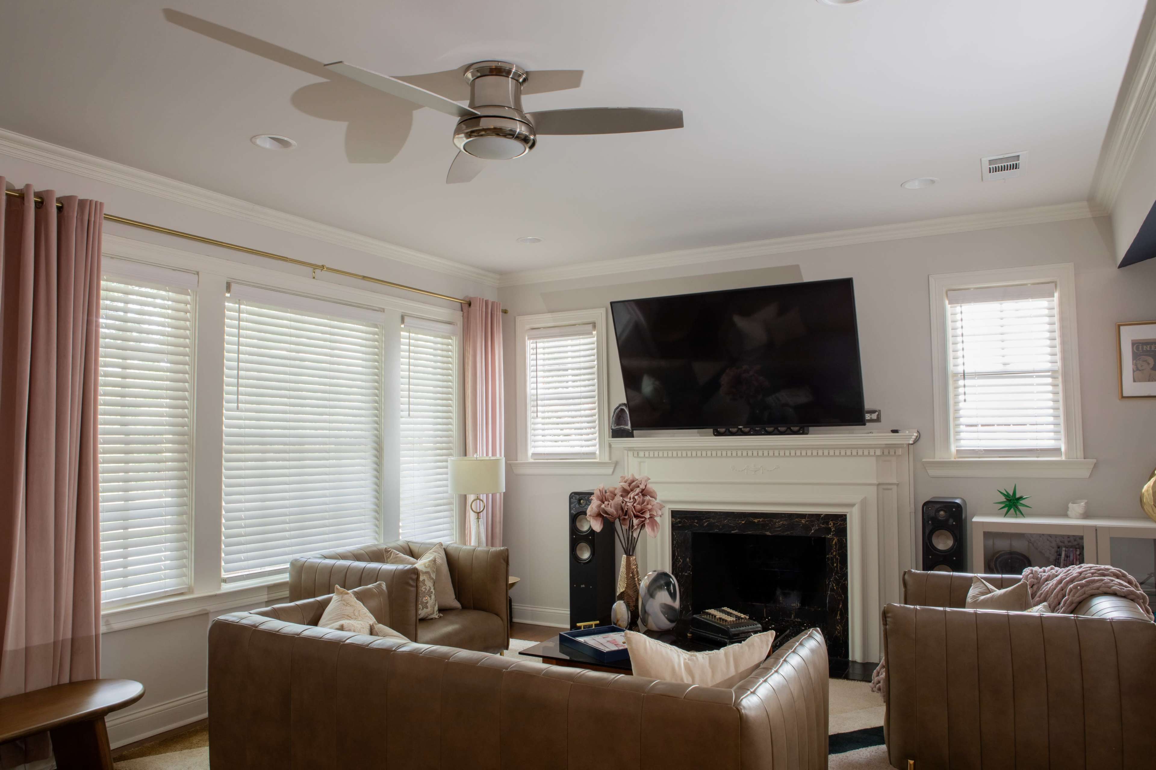 The image shows a living room with two leather sofas, a television mounted above a fireplace, and large windows covered with blinds.