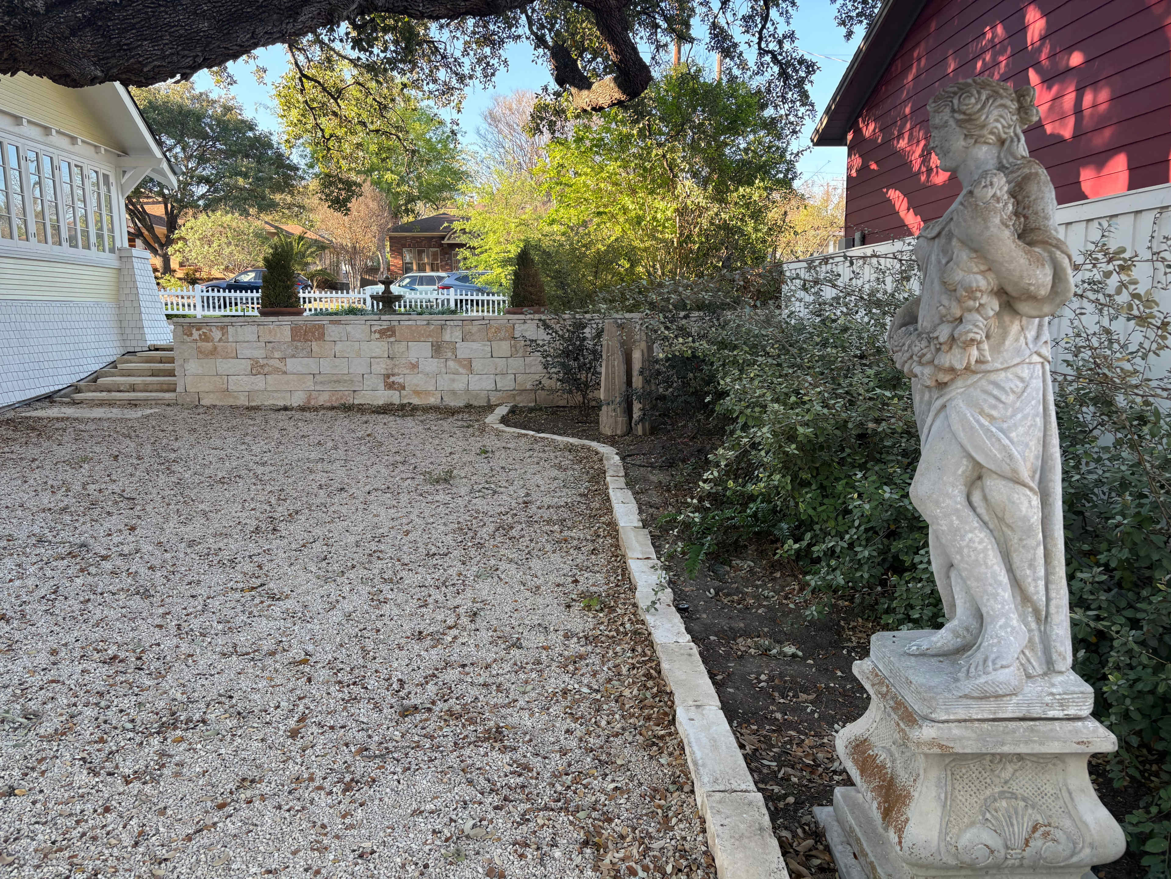 A stone statue of a figure holding a small animal stands beside a gravel path that curves along a landscaped area near a red building.