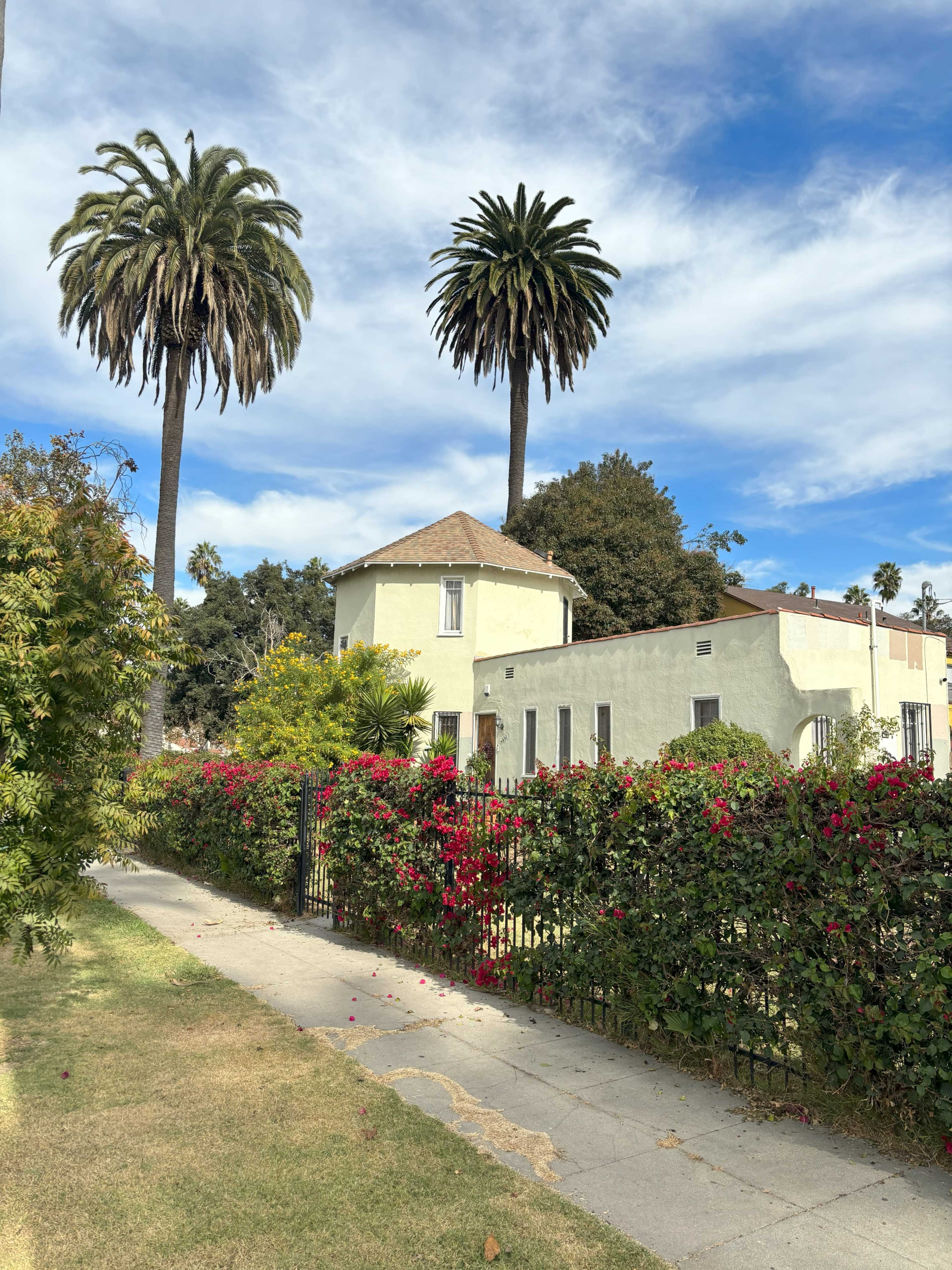 A pale yellow house with a round shape is surrounded by lush greenery and tall palm trees under a blue sky.