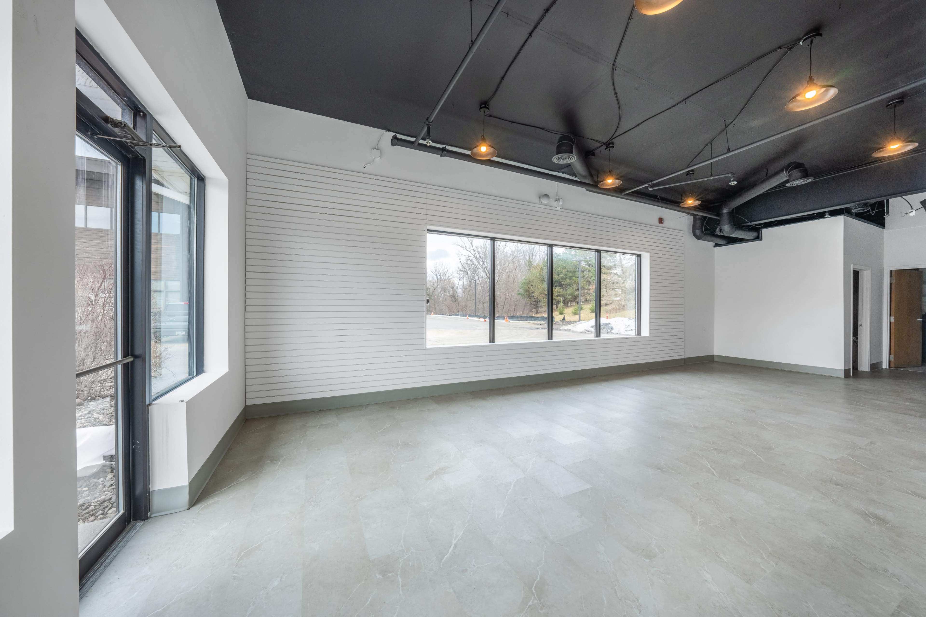 The image shows an empty interior space with large windows allowing natural light, a smooth floor, and exposed ceiling fixtures.