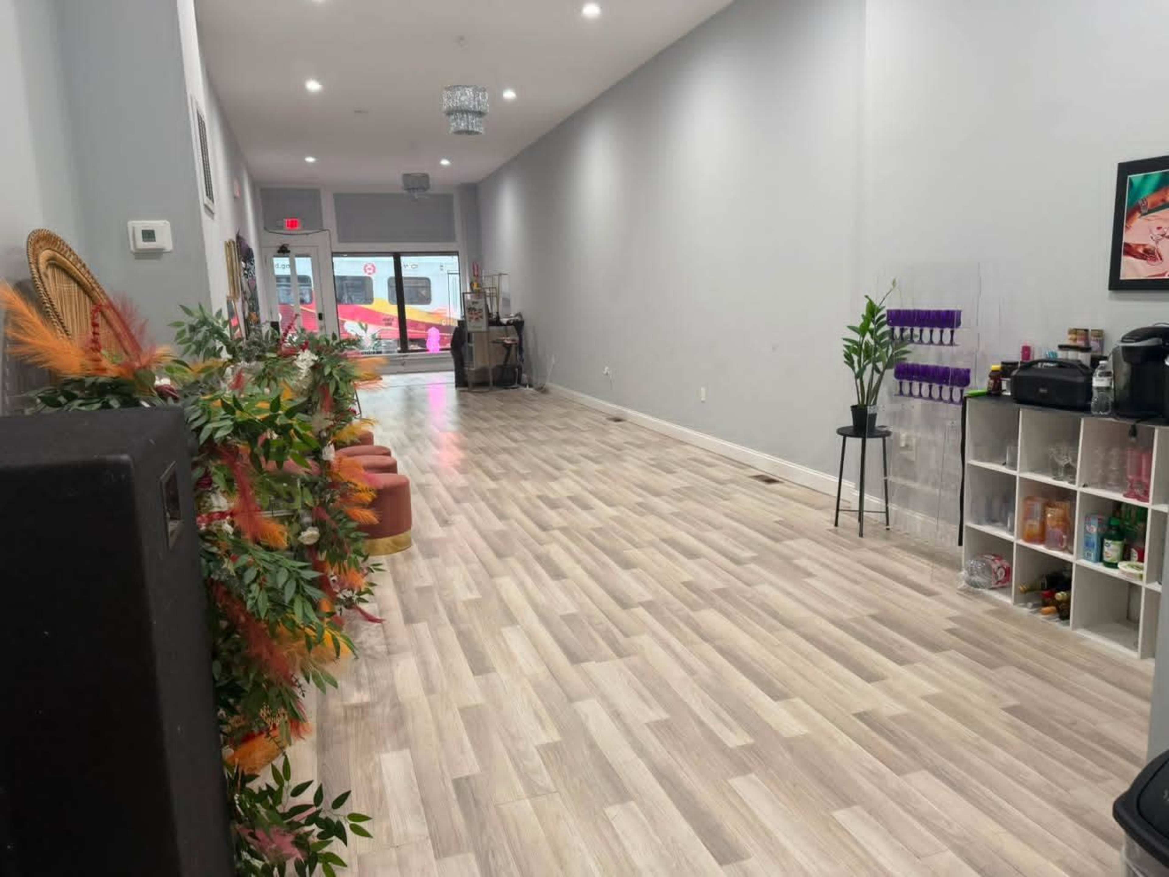 The image shows a narrow, well-lit indoor space with light-colored flooring, a row of plants on one side, and furniture arranged along the walls.