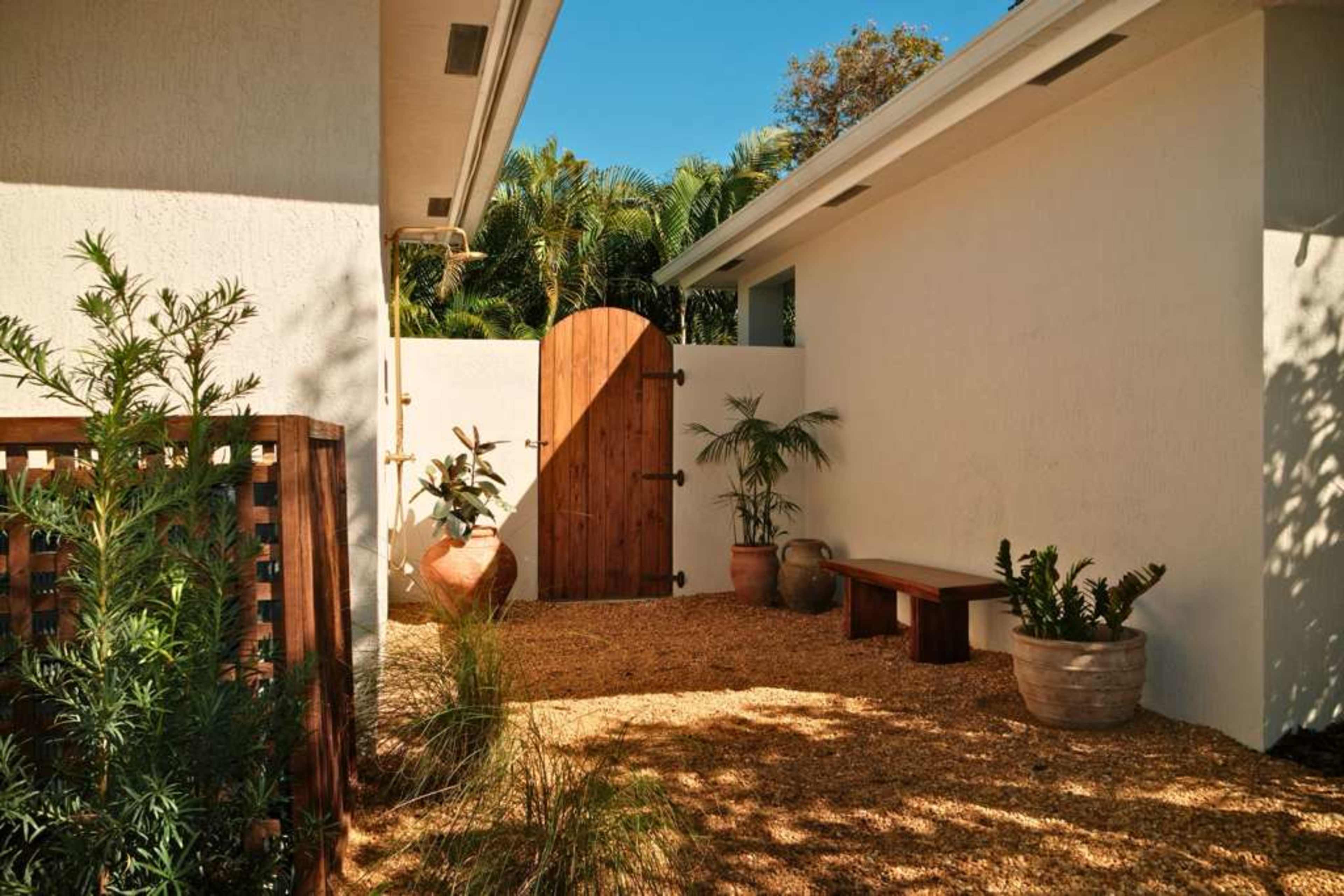 The image shows a small, enclosed courtyard featuring a wooden gate, a stone bench, and several potted plants surrounded by gravel.