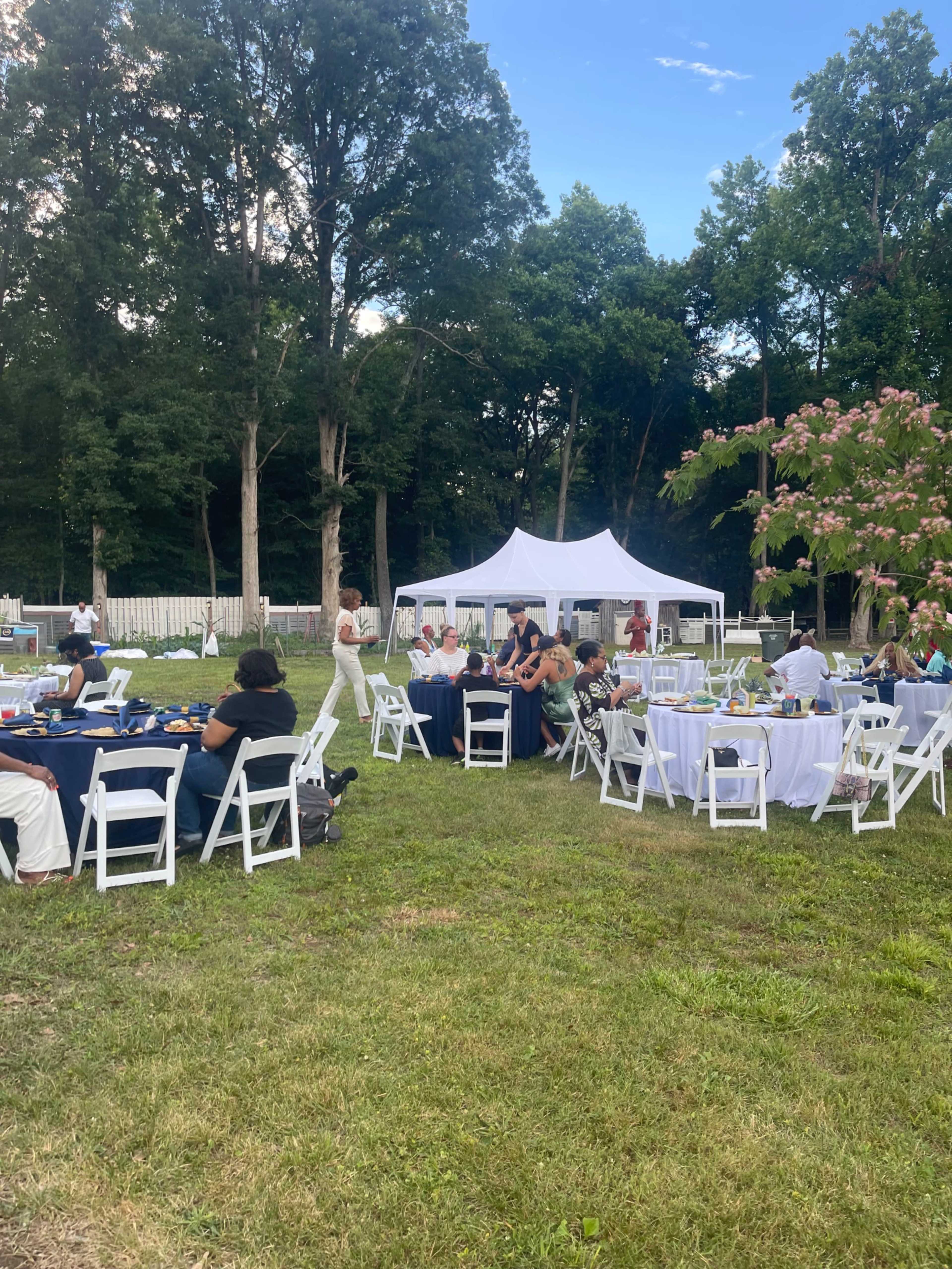 A gathering is taking place outdoors with seating arrangements at tables and a large white tent in the background.