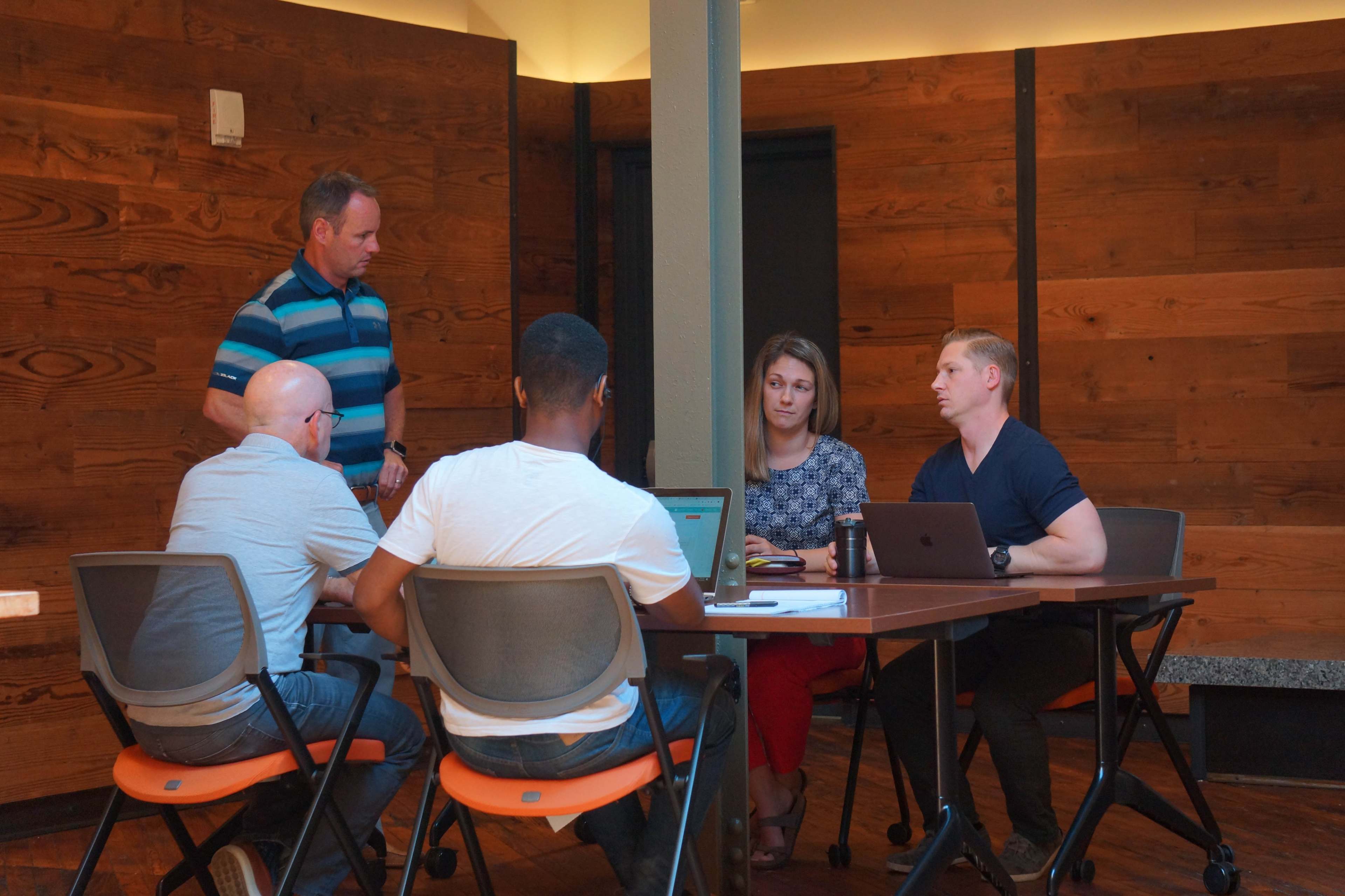 A group of four people is seated around a table in a conference room discussing a project, while another person stands nearby.