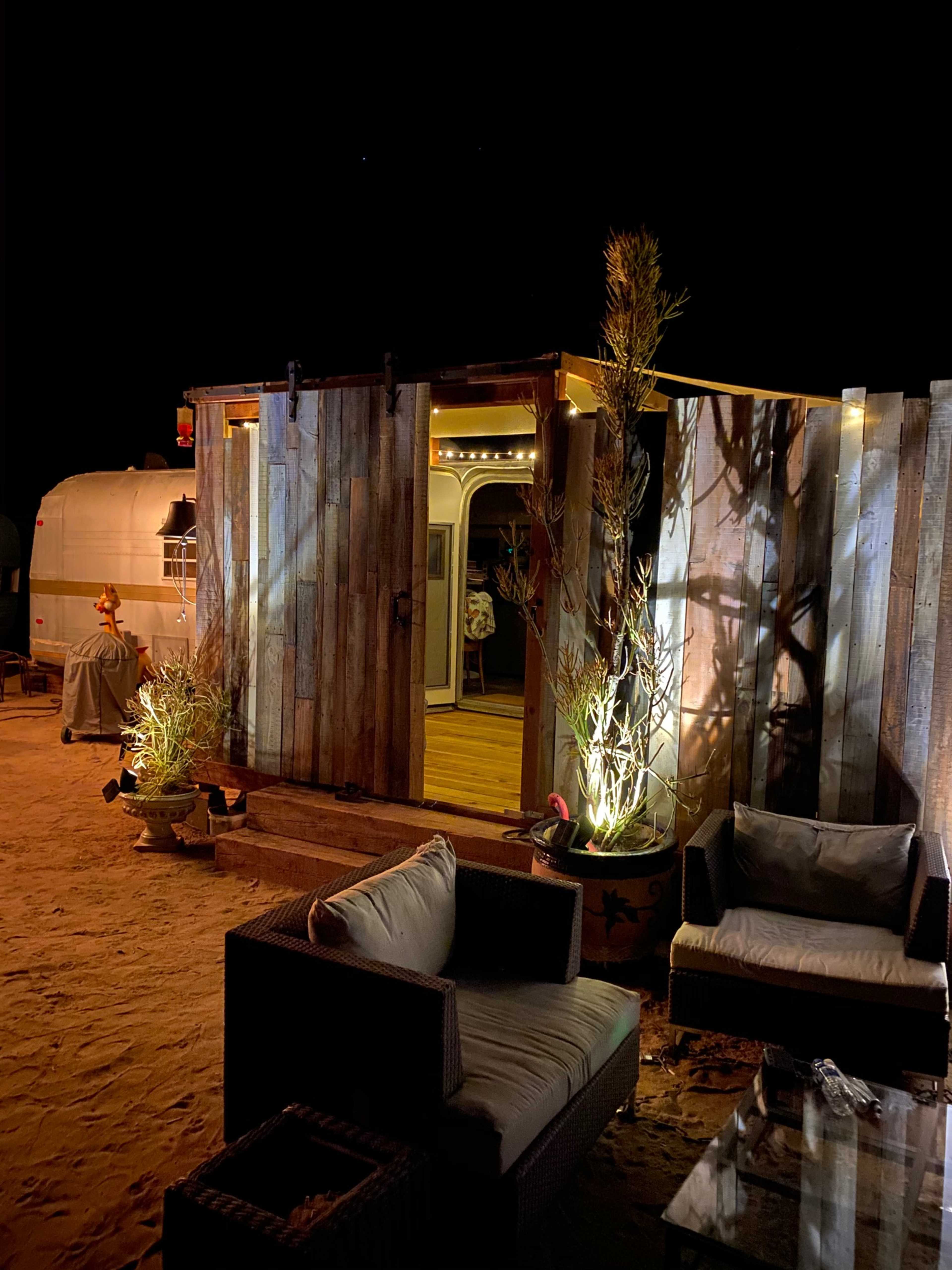 A wooden structure with a lit entrance is surrounded by sandy ground and illuminated plants at night.