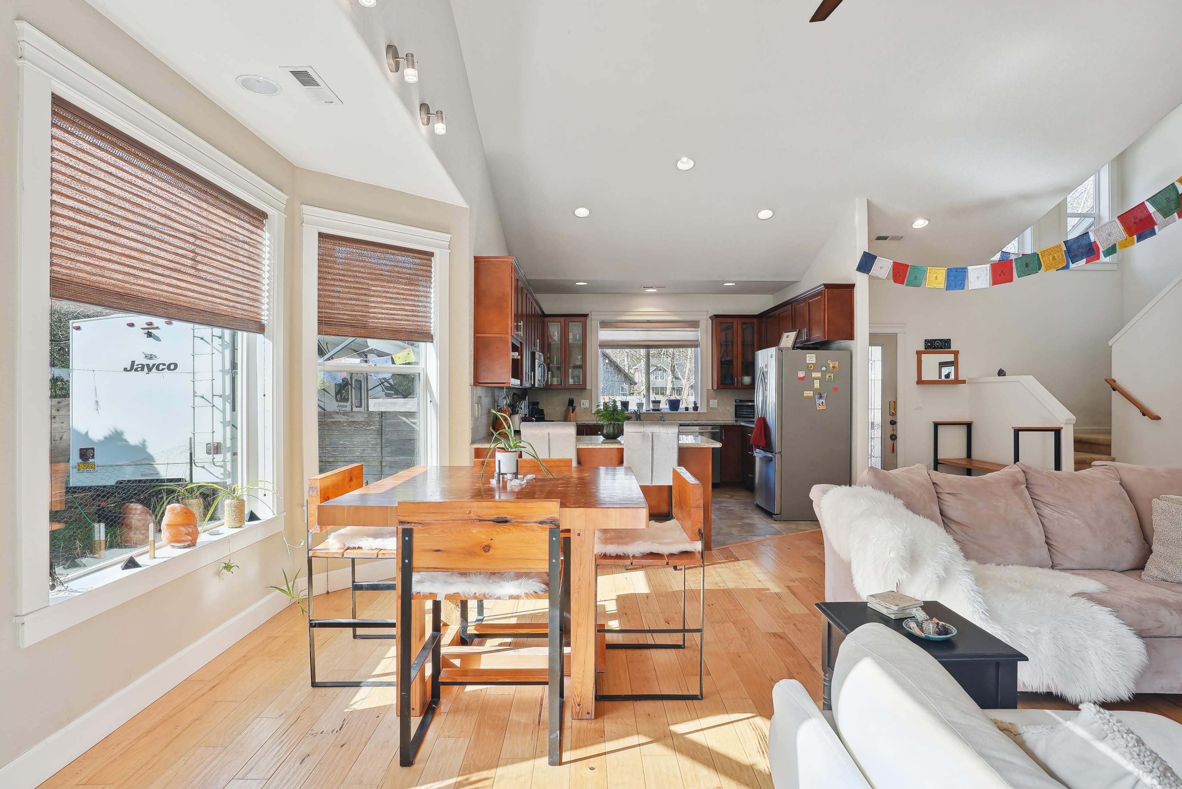 A modern living space features a dining area with a wooden table and chairs, surrounded by large windows, open to a kitchen that includes a refrigerator and wood cabinets.