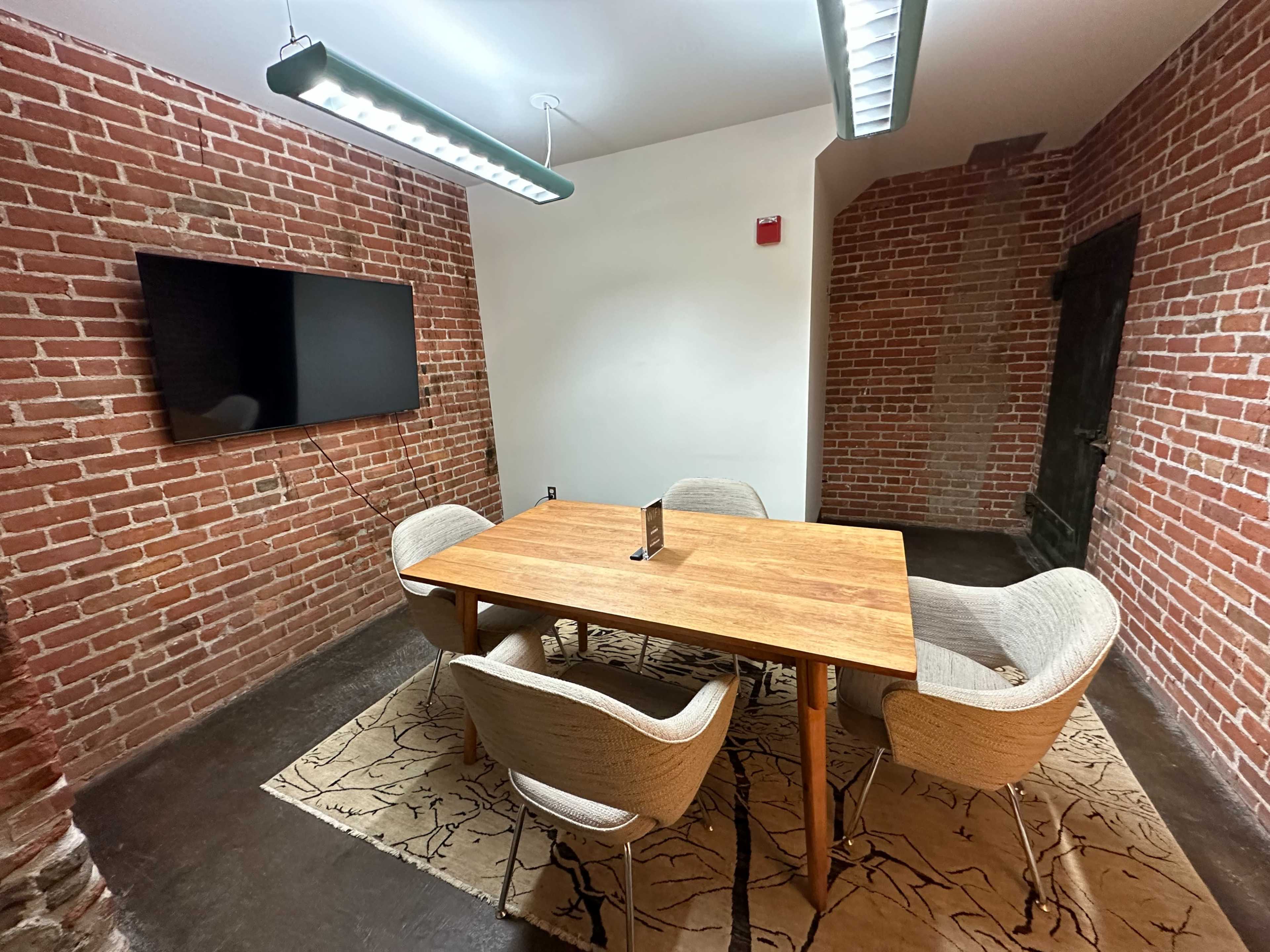A small meeting room features a wooden table surrounded by four upholstered chairs, a wall-mounted television, and exposed brick walls.