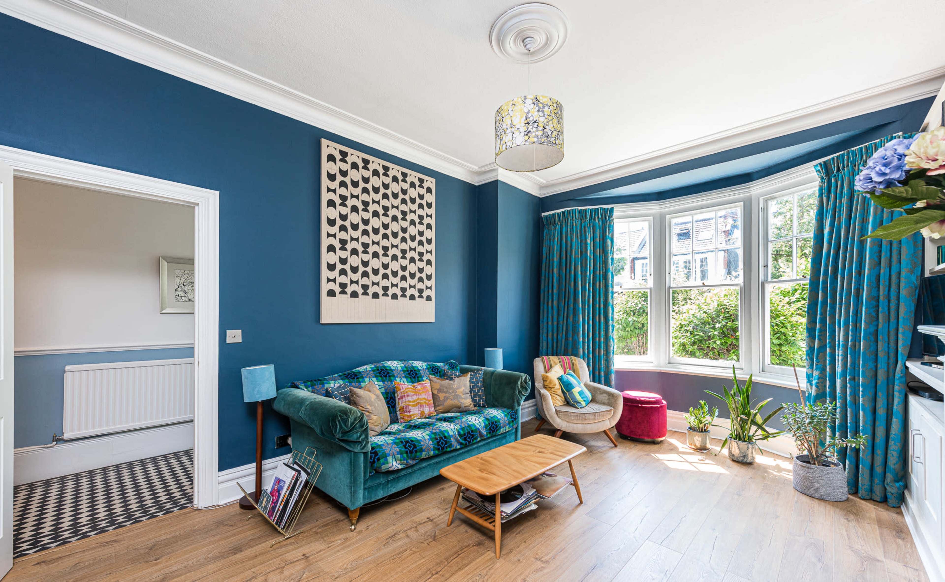 A bright living room features a blue accent wall, a patterned artwork above a green sofa, and a large bay window with plants nearby.