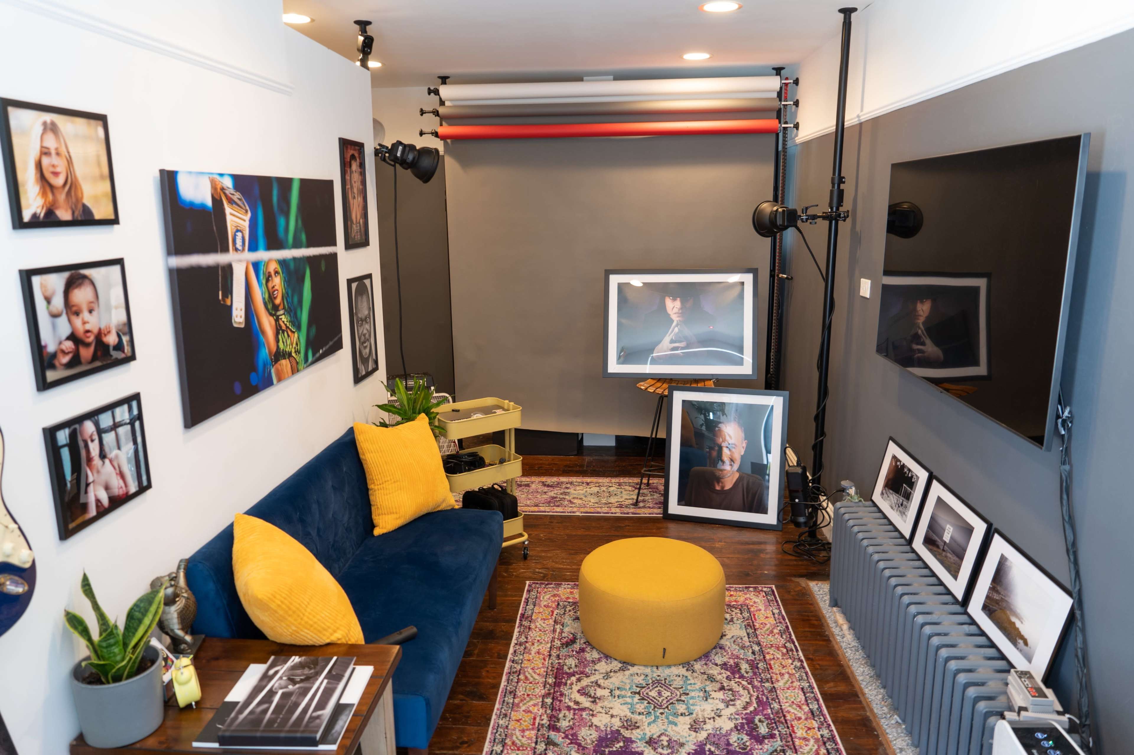A styled photo studio featuring a blue sofa with a yellow cushion, framed photographs on the walls, and a circular ottoman on a patterned rug.