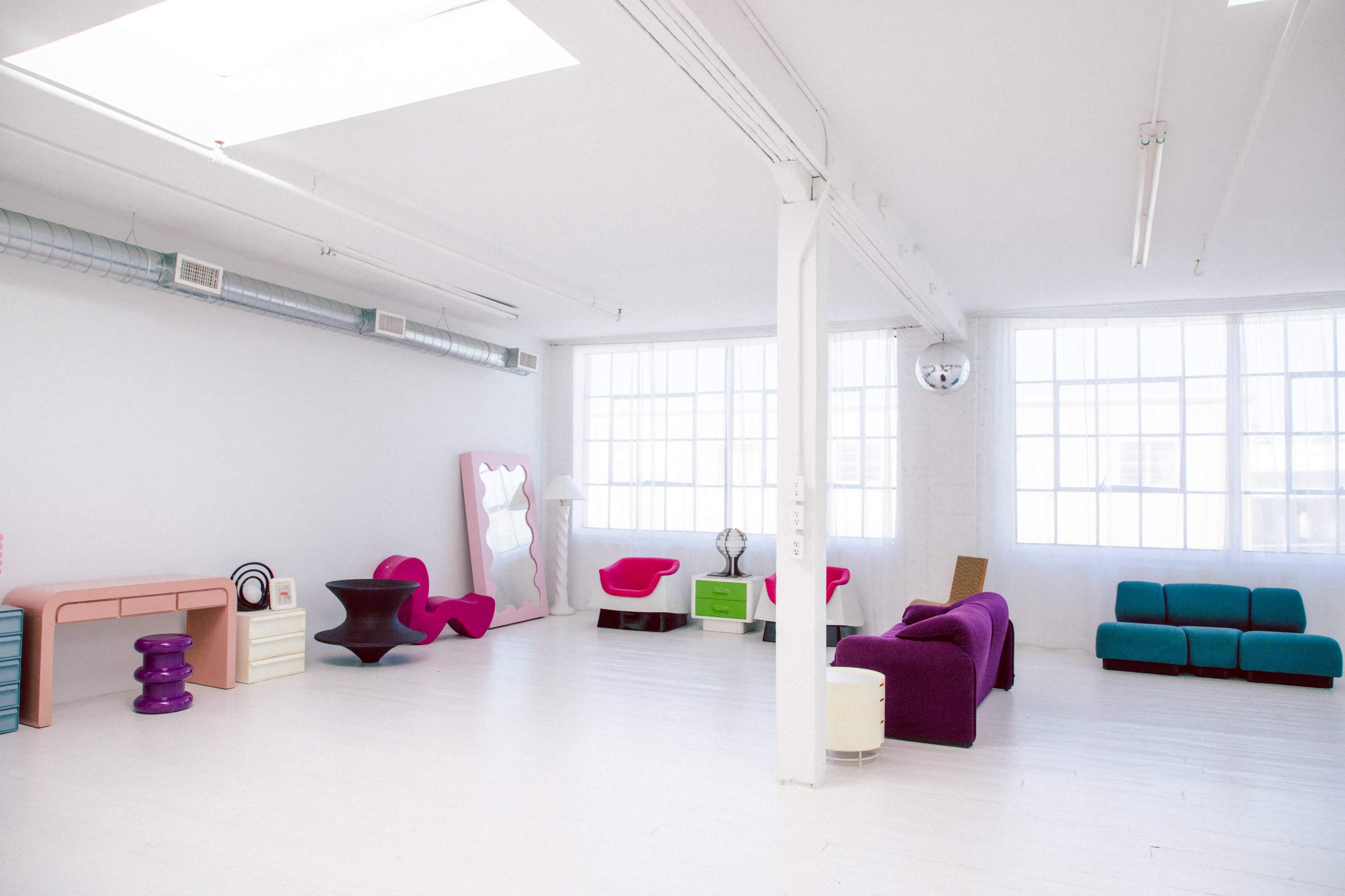 A spacious, well-lit room featuring colorful modern furniture, including a pink sofa, teal seating, and a unique purple table, against a backdrop of white walls and large windows.